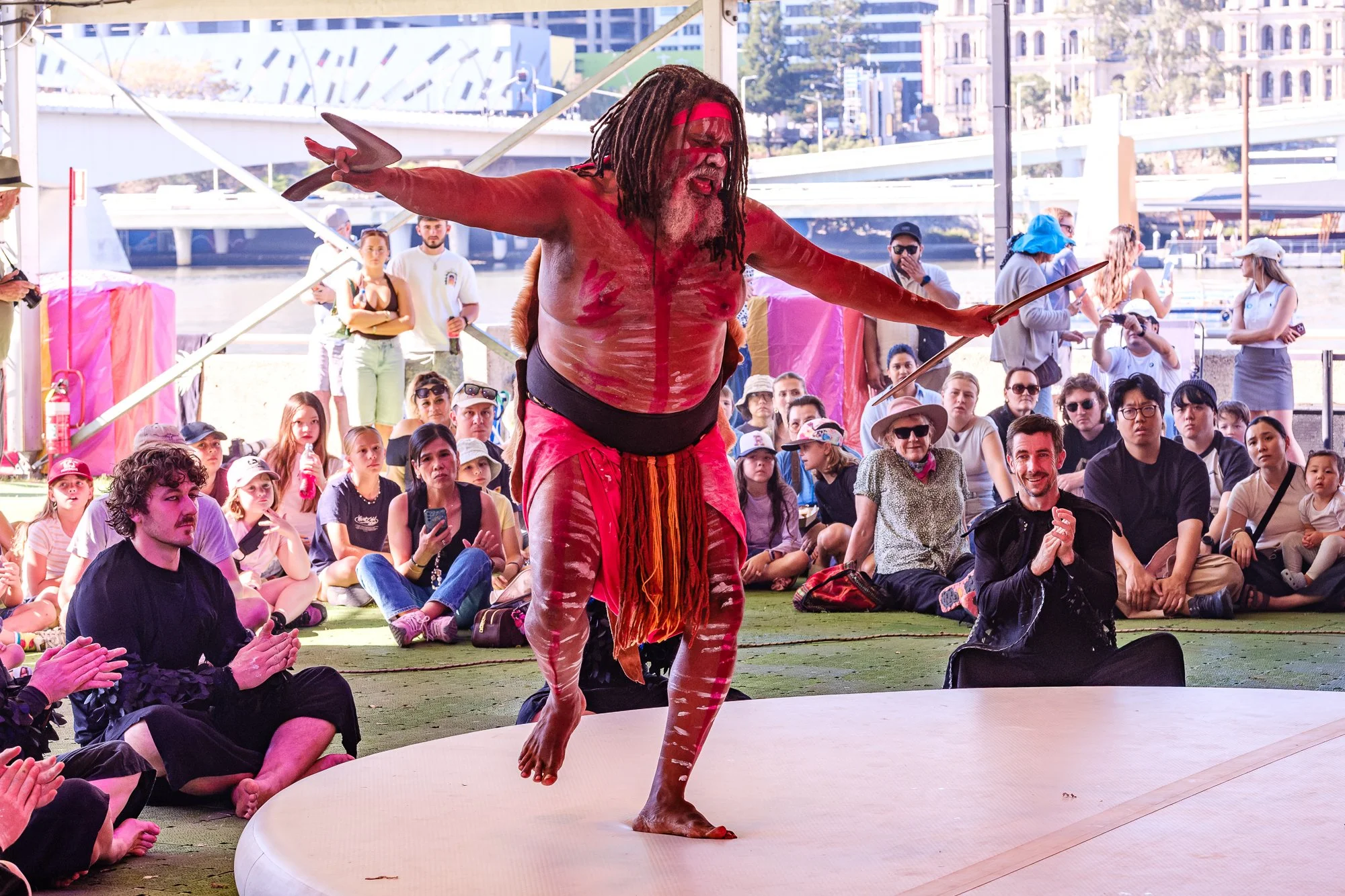 A performer dressed in traditional tribal attire with painted body, dreadlocks, and face paint, is dancing barefoot on a raised platform during a cultural show. The audience, sitting on the ground, watches attentively, with some clapping and others c