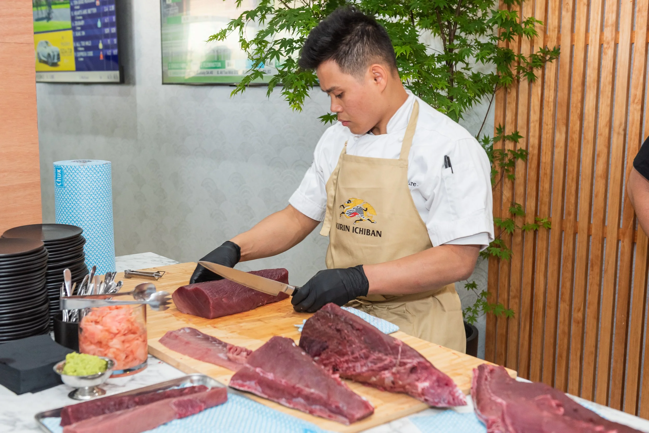 Chef slicing raw beef at a cutting board with other cuts of beef nearby, in a modern setting with a wood and greenery background.