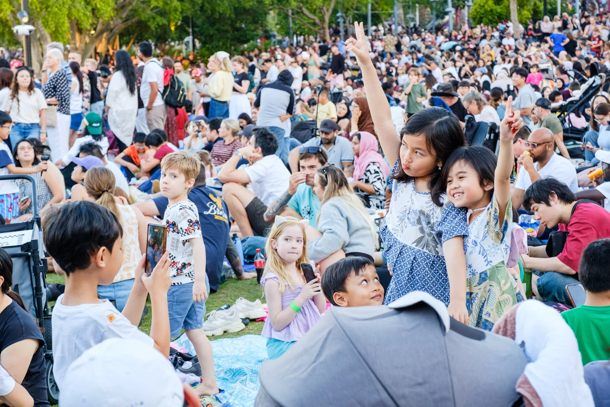 A large crowd of diverse people gathered outdoors in a park with many sitting on the grass and some standing. A young woman is raising her hand, and others are engaging in conversations or taking photos.