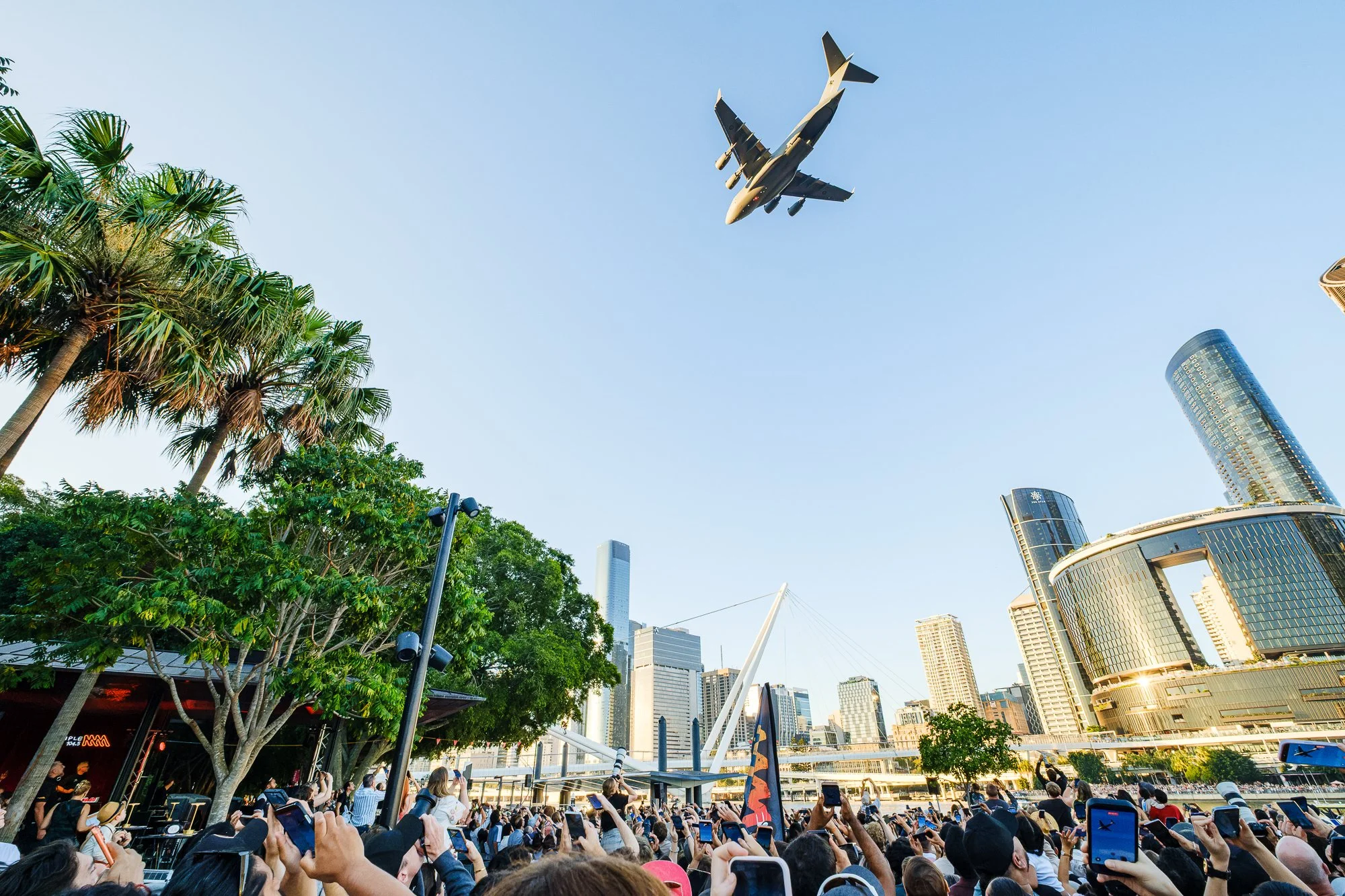 A large crowd of people taking photos and videos with their smartphones in front of a skyline with modern skyscrapers, palm trees, and a fighter jet flying overhead in clear blue sky.