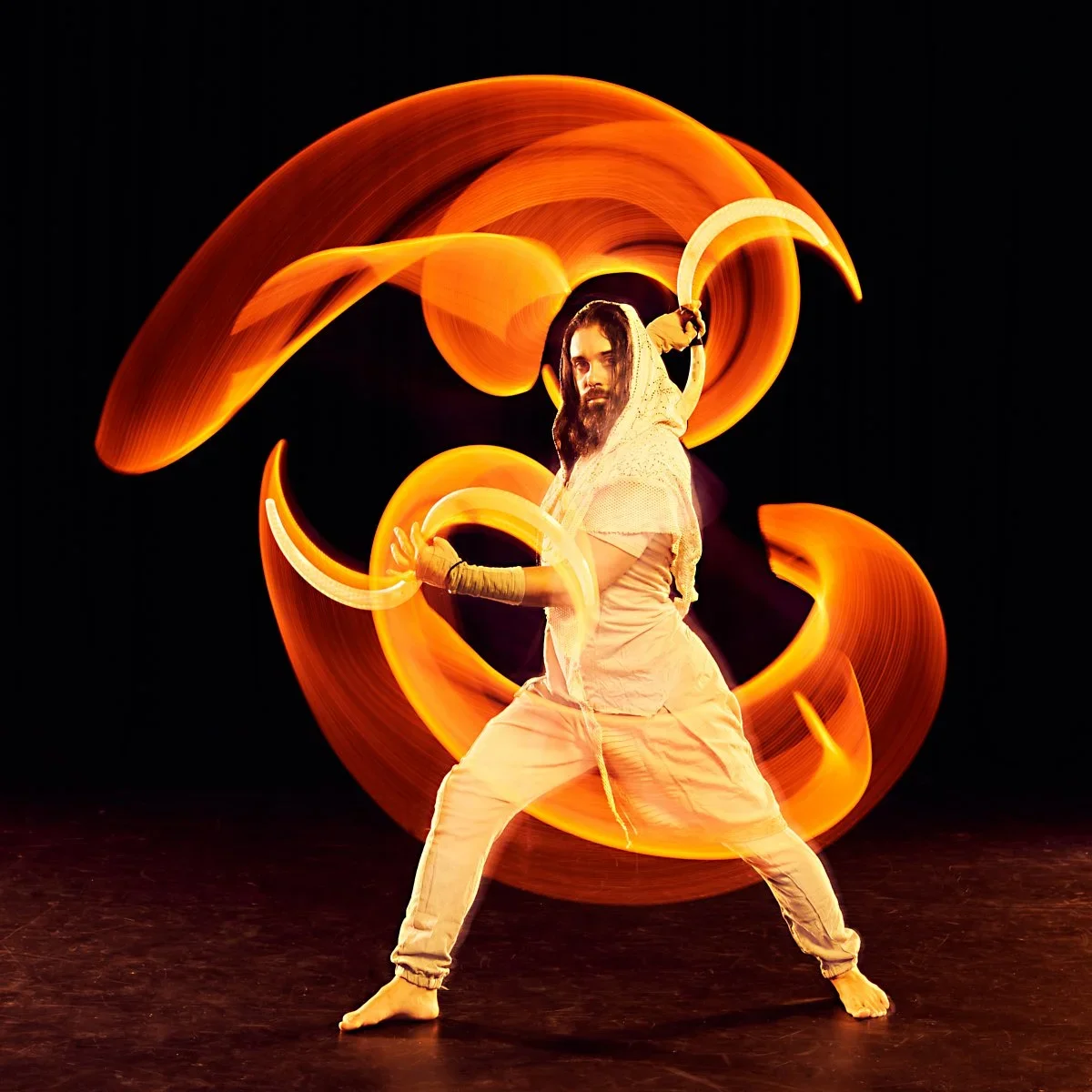 A person dressed in beige performing with fire poi against a black background, creating swirling orange flame trails.