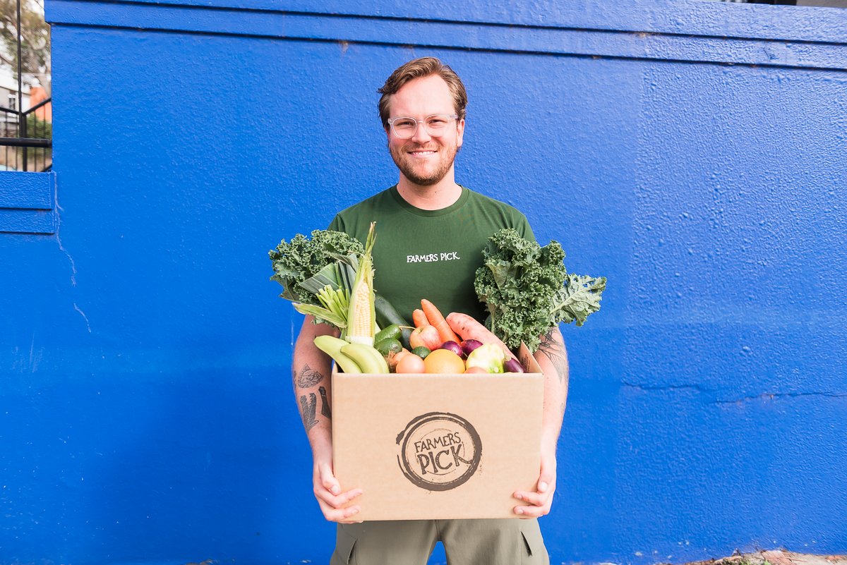 A man with glasses and a beard holding a box of fresh vegetables, standing in front of a bright blue wall.