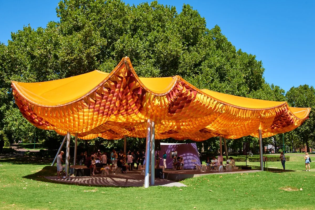 Colorful outdoor pavilion with yellow and orange canopy, in a park with green grass and large trees, and people gathering underneath.