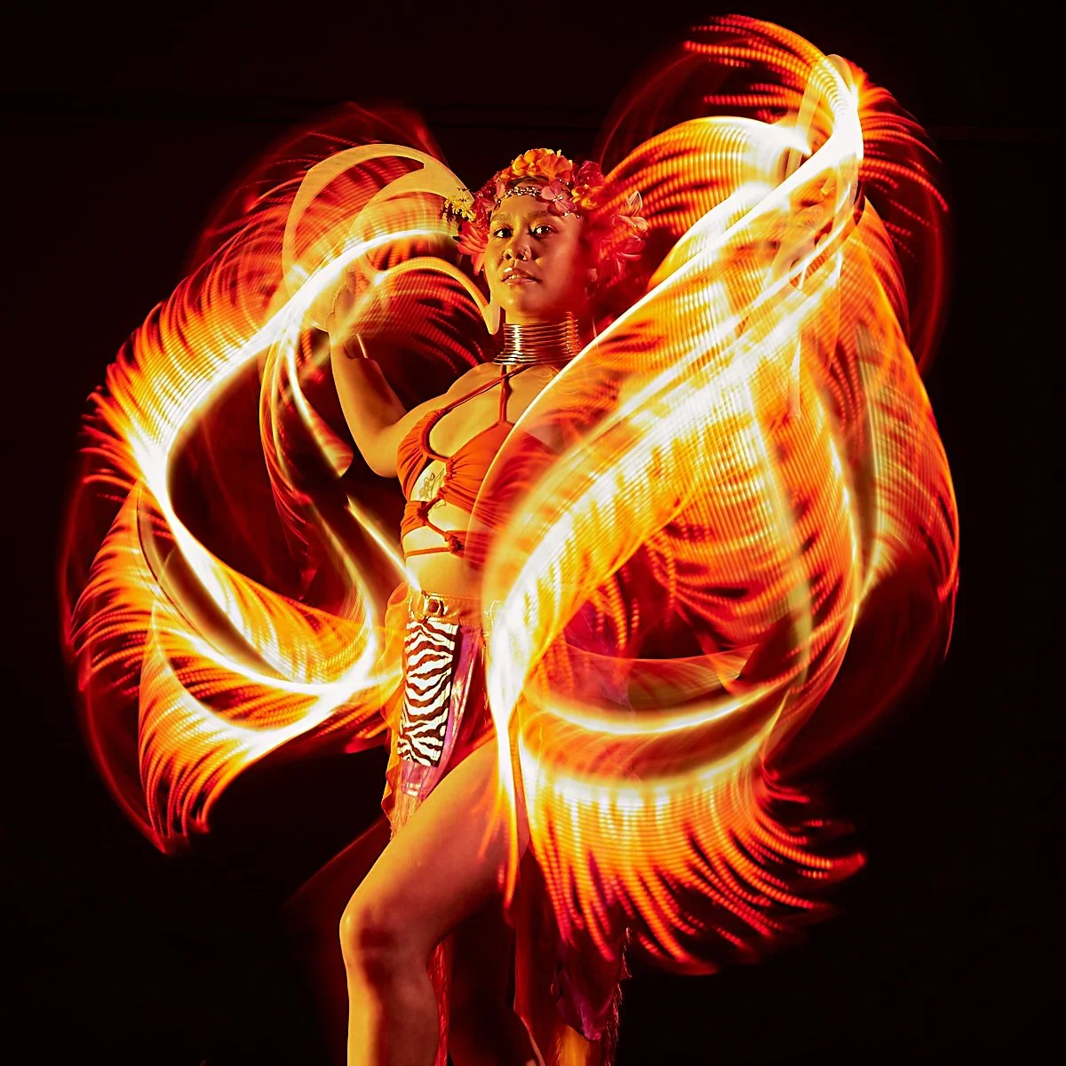 A woman in colorful costume with fire-like light trails swirling around her, creating a dynamic and vibrant visual effect.