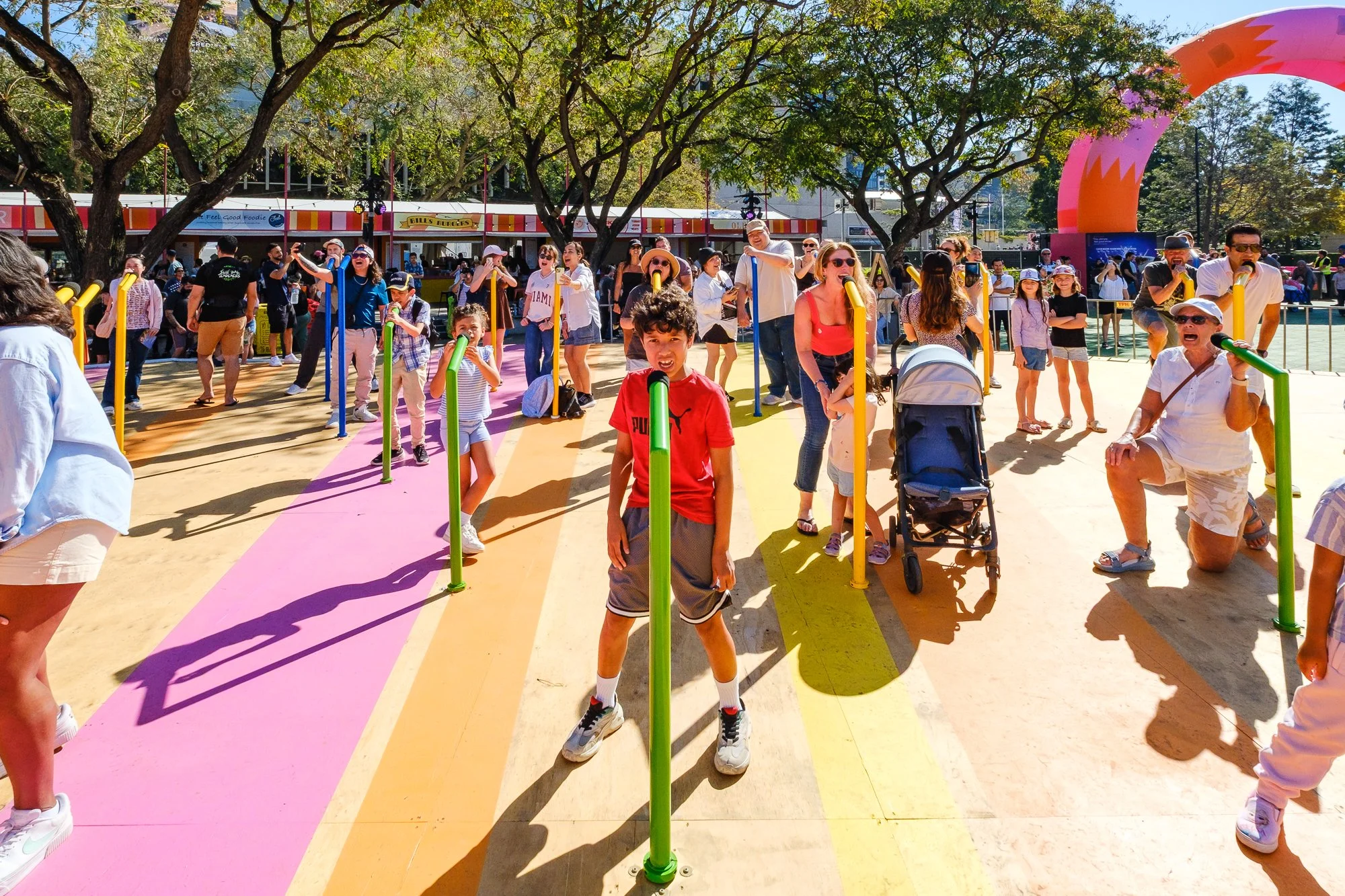 Children and adults participating in a colorful outdoor activity involving a rainbow-colored pathway and a pink dinosaur arch. Some people are taking photos and playing with yellow and green plastic tube toys, with trees and food stalls in the backgr