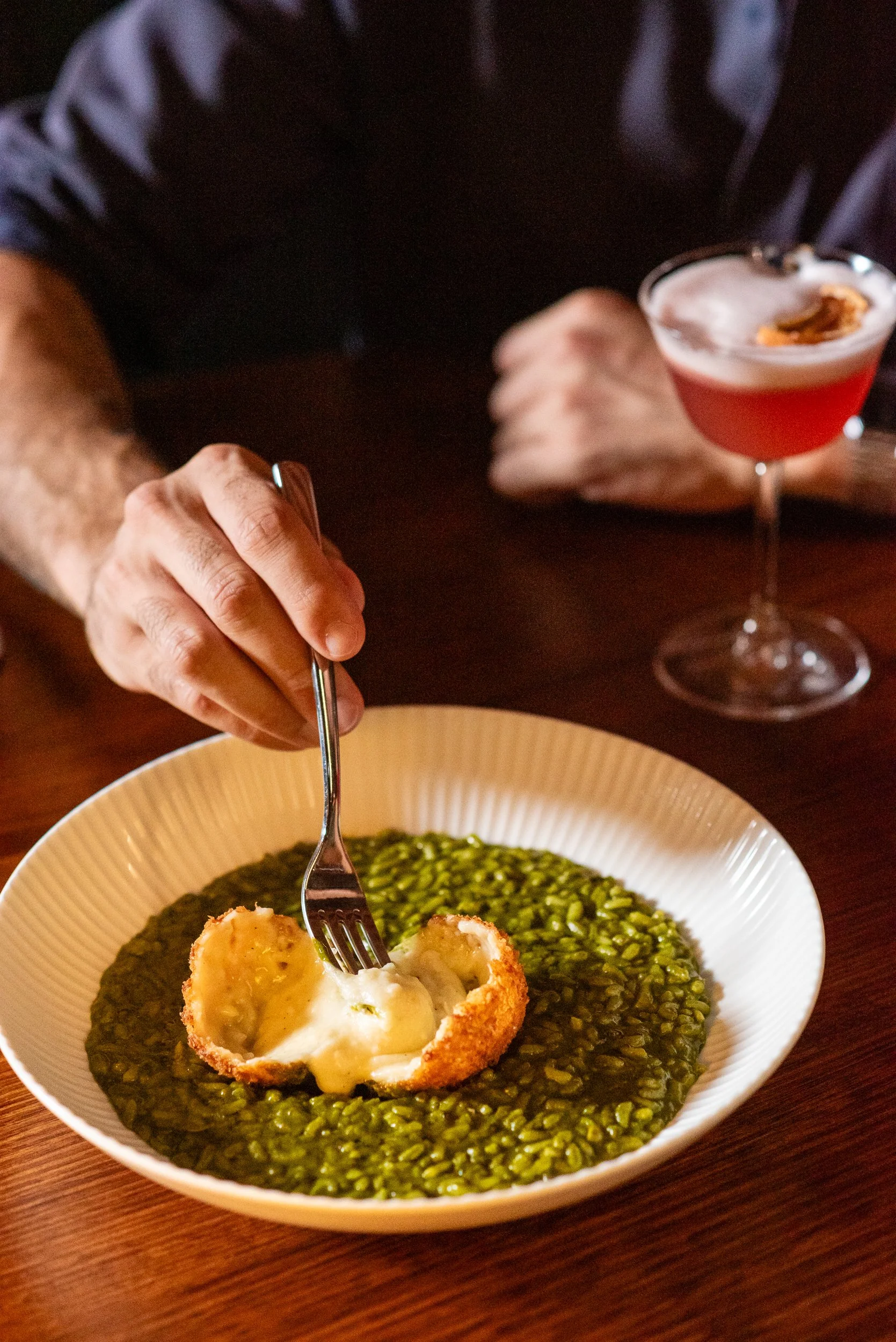 A person holding a fork over a white bowl of green lentil soup with a breaded, baked eggplant topped with melted cheese and a scoop of creamy white sauce on top, and a woman holding a cocktail in a glass with a pinkish hue and a slice of fruit garnis