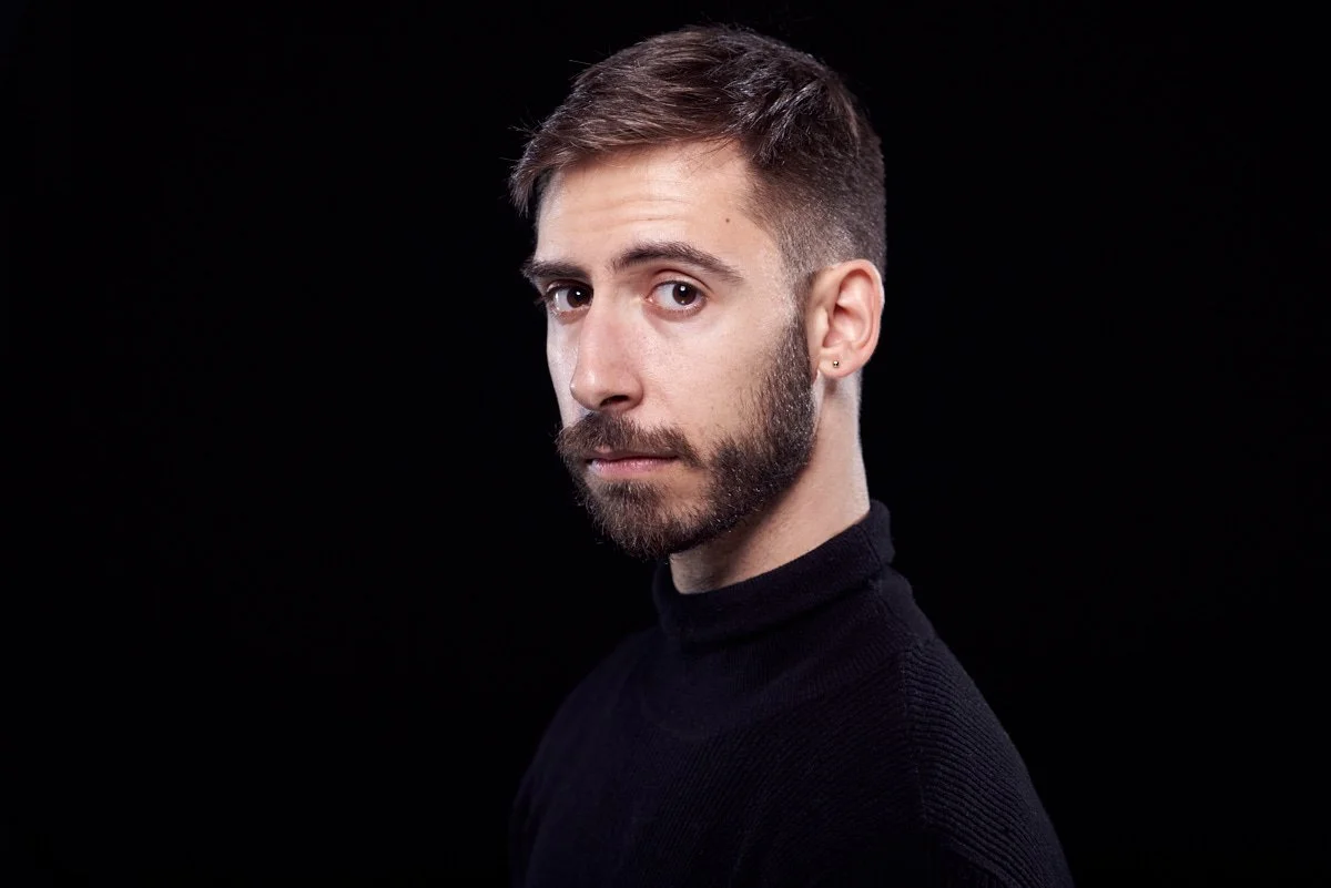 Portrait of a young man with short brown hair, a beard, and earrings, wearing a black turtleneck, against a black background.
