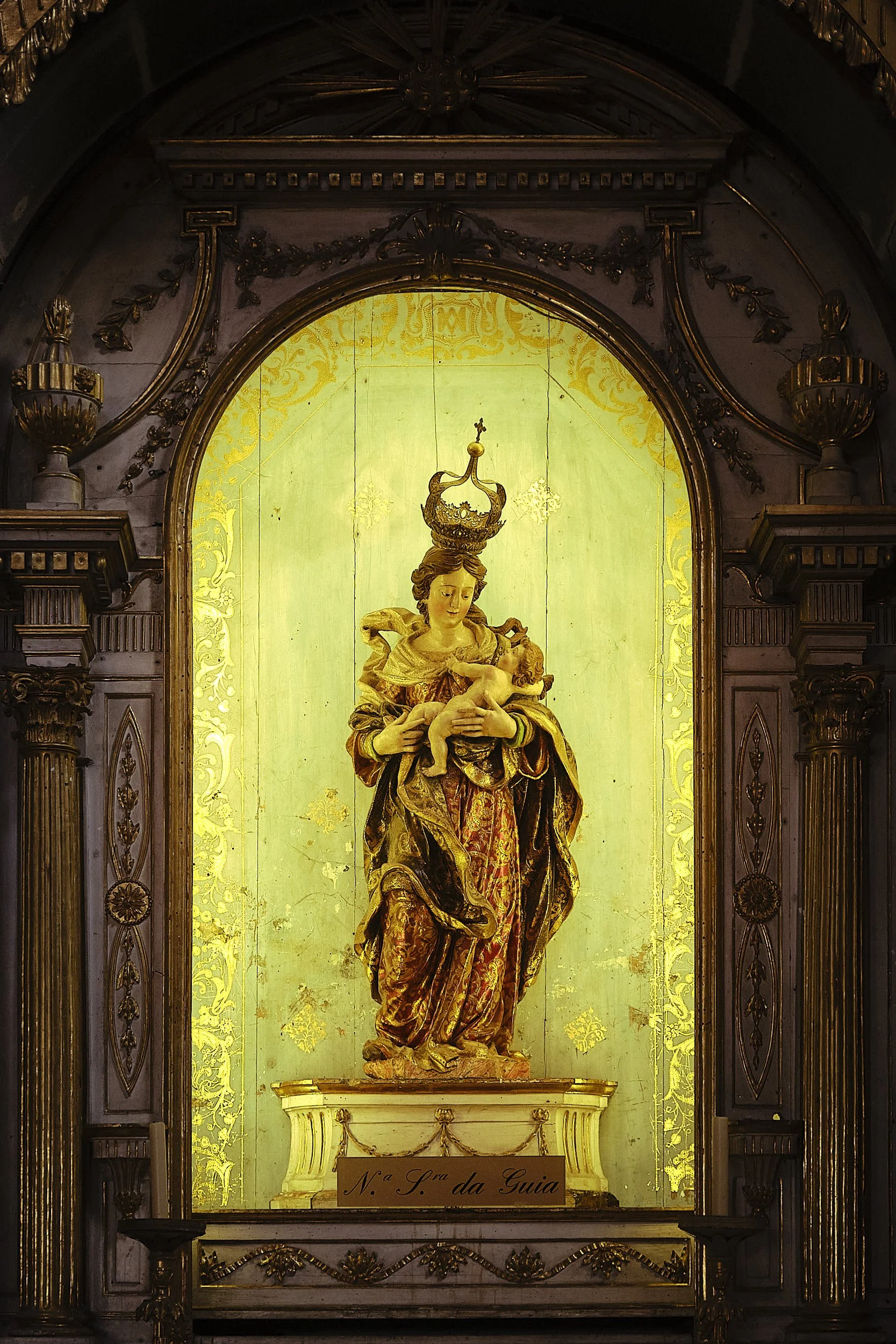 A religious statue of the Virgin Mary holding baby Jesus, adorned with a crown, in a decorated church altar with gold accents and an illuminated backdrop.