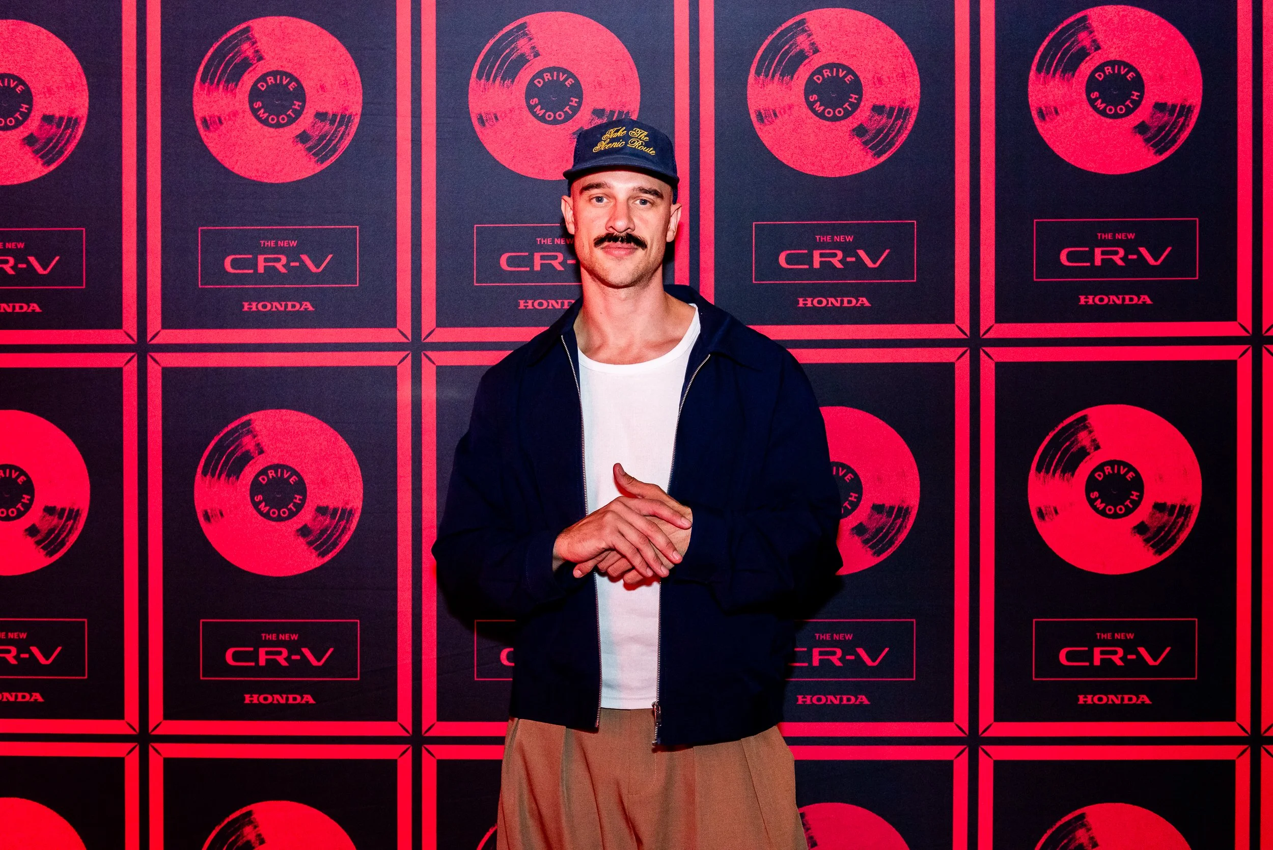 A man with a mustache wearing a navy jacket, white t-shirt, beige pants, and a navy cap with gold embroidery, standing in front of a red and black backdrop with repeated graphics of a vinyl record and the words "The New CR-V" and "Honda."