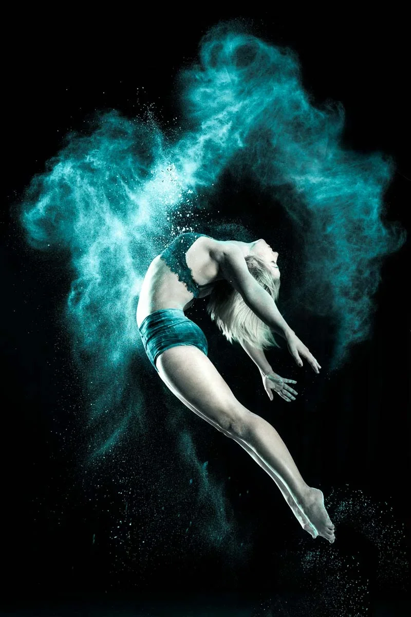 A woman performing underwater dance or acrobatics surrounded by a cloud of blue powder or dust against a black background.