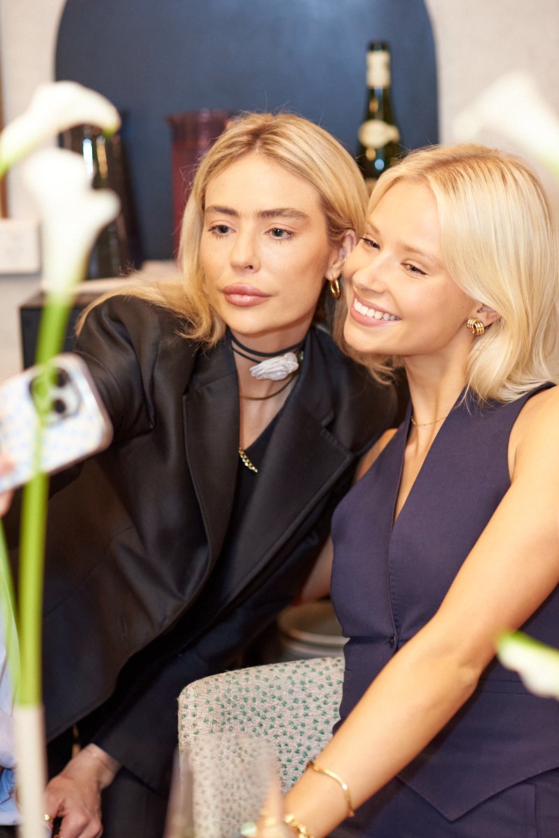 Two women taking a selfie together at a social gathering, with flowers and bottles in the background.
