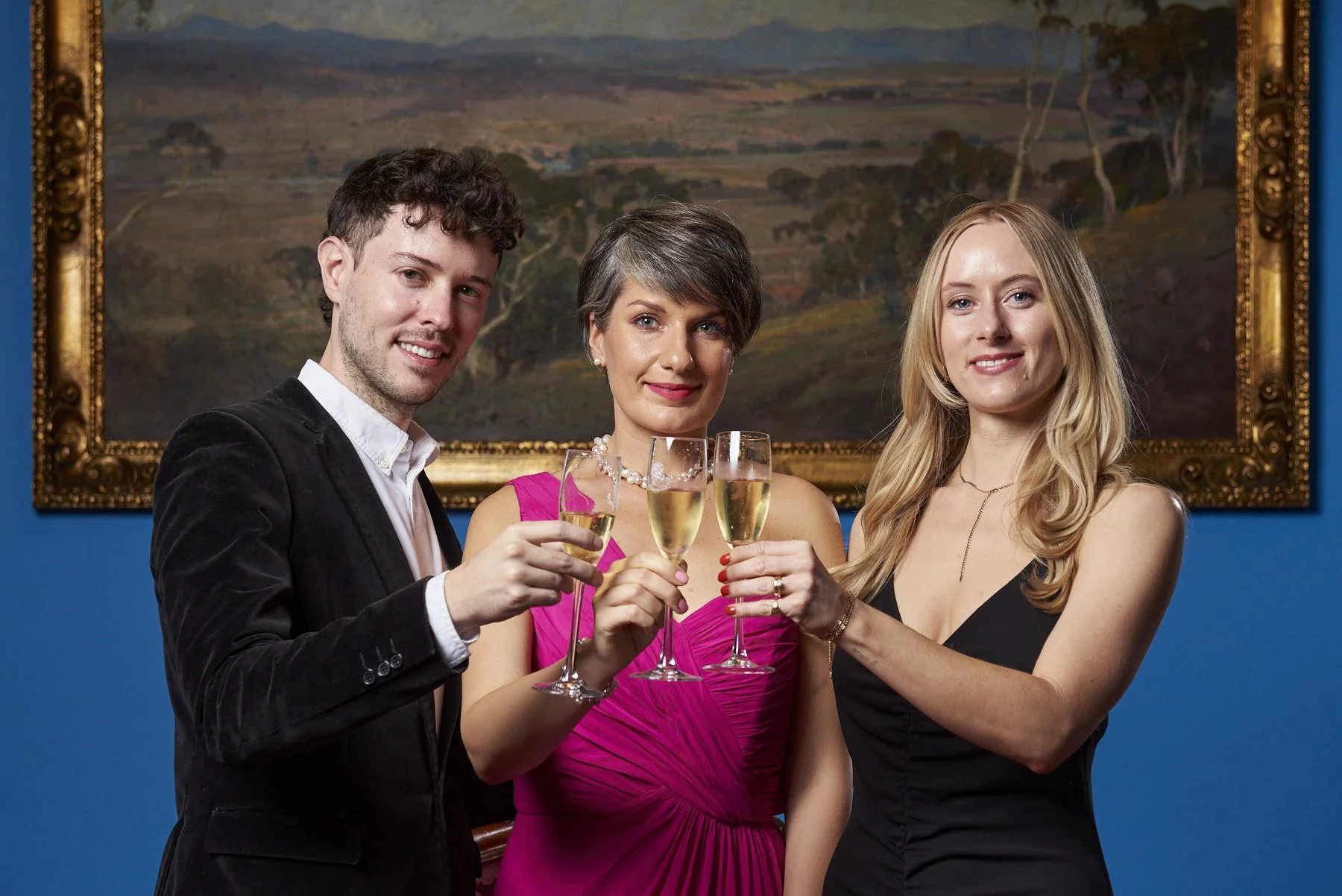 Three people celebrating with champagne glasses in front of a landscape painting on a blue wall.