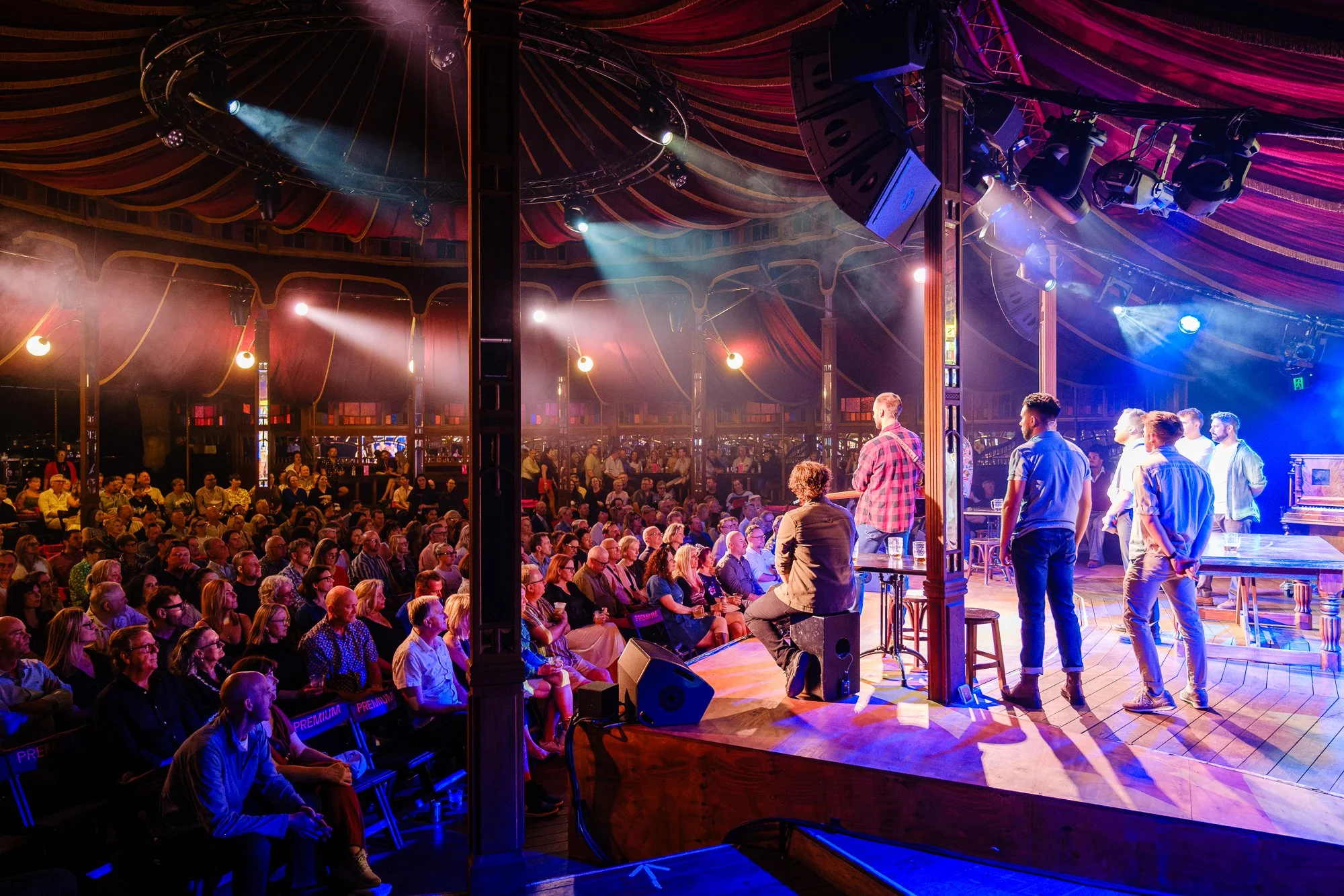 Audience watching a performance on stage in a theater with red curtains and stage lighting.