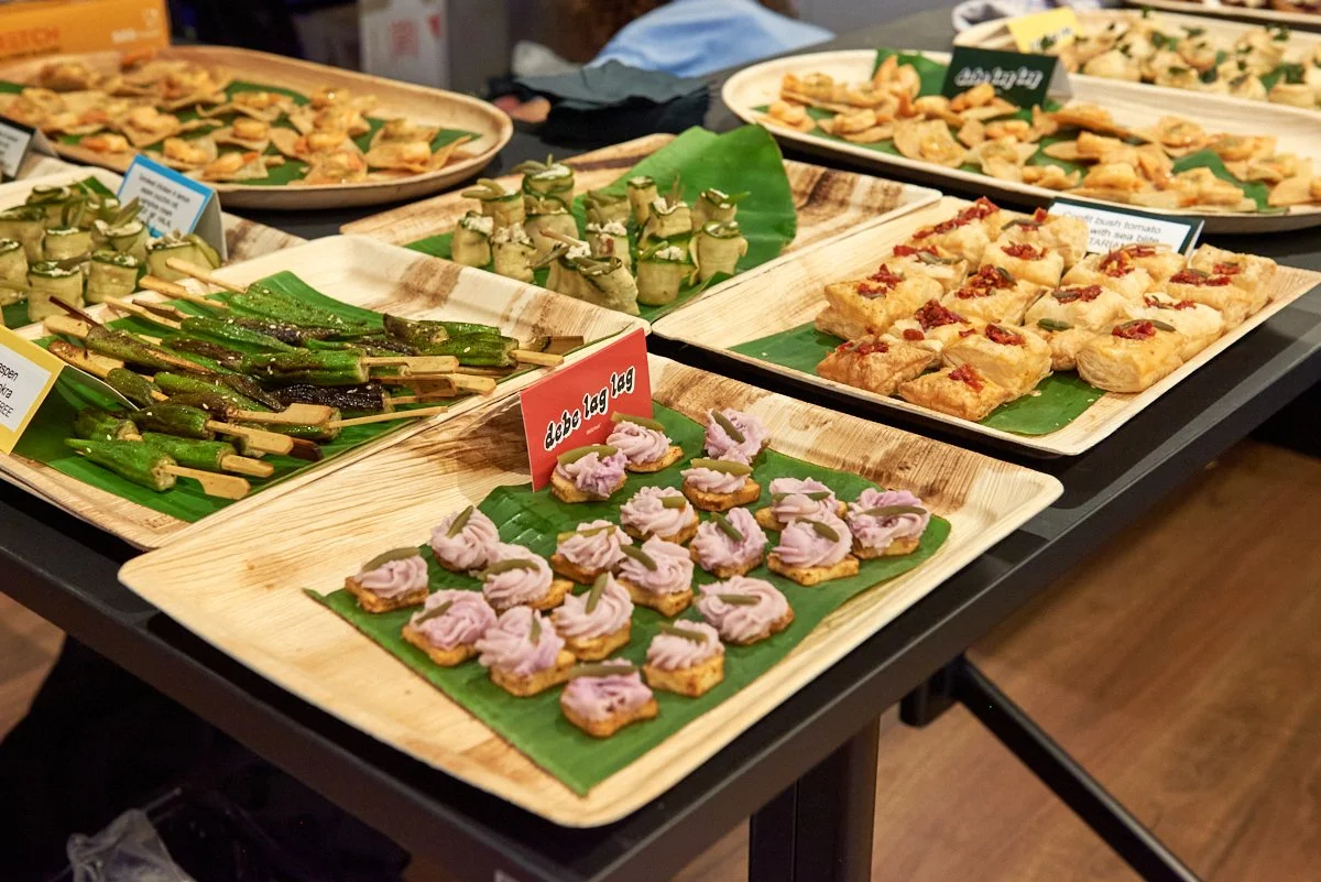 Assorted appetizers on wooden trays with banana leaf liners, including skewered green vegetables, cucumber rolls, small savory pastries, and crackers topped with pink and red spreads at a buffet table.
