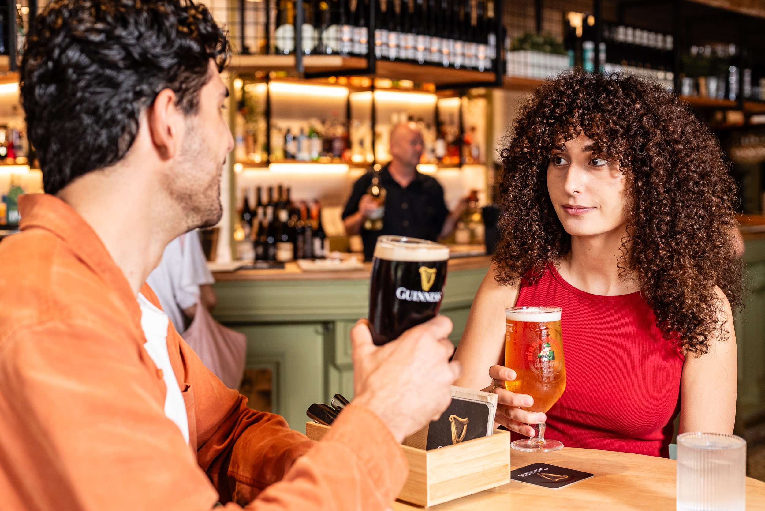 A man and a woman seated in a bar, holding glasses of dark stout and orange ale, having a conversation. The woman has curly hair and is wearing a red top, the man has short curly hair and is wearing an orange jacket.