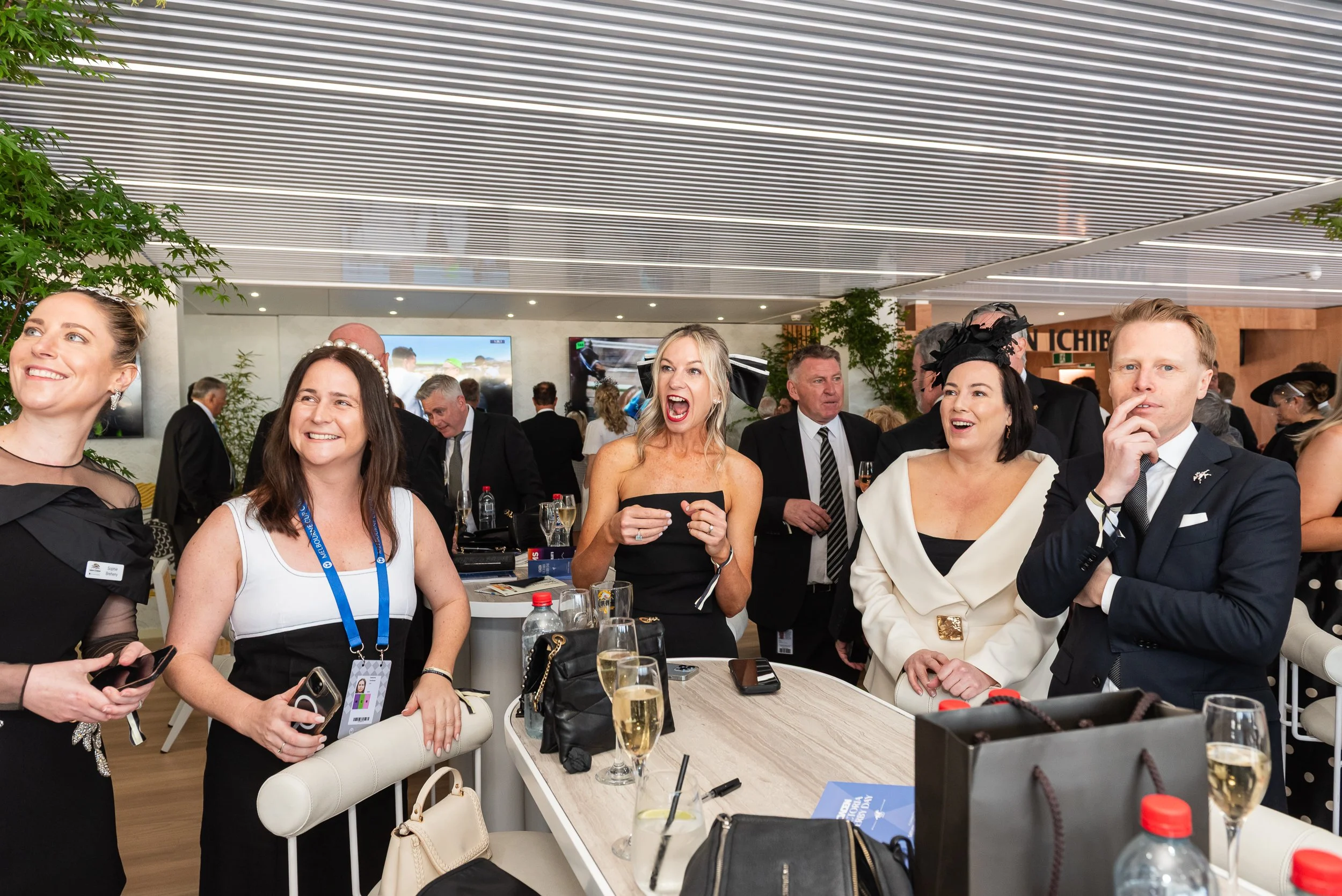 Group of people at a social event, dressed in formal attire, in a modern indoor setting with a ceiling with linear lighting, drinks on the table, and background screens.