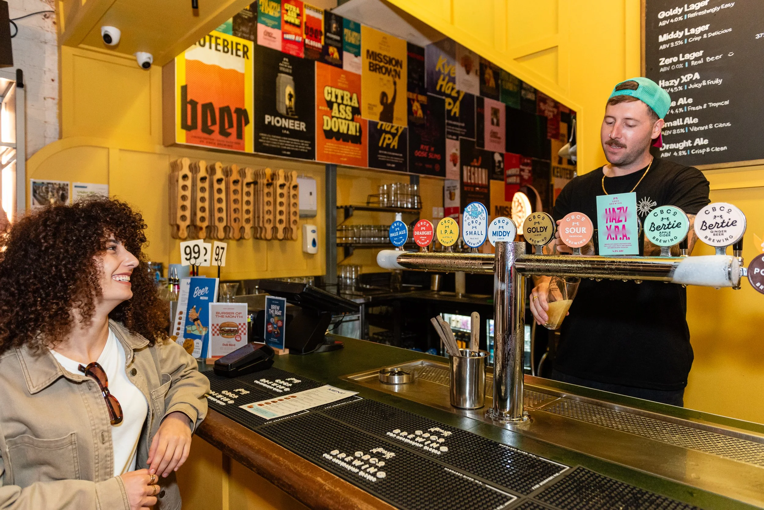 A woman with curly hair and sunglasses on her head smiling at a bartender with a mustache, wearing a teal backward cap and black shirt, at a craft brewery or bar with a bright yellow wall and various posters.