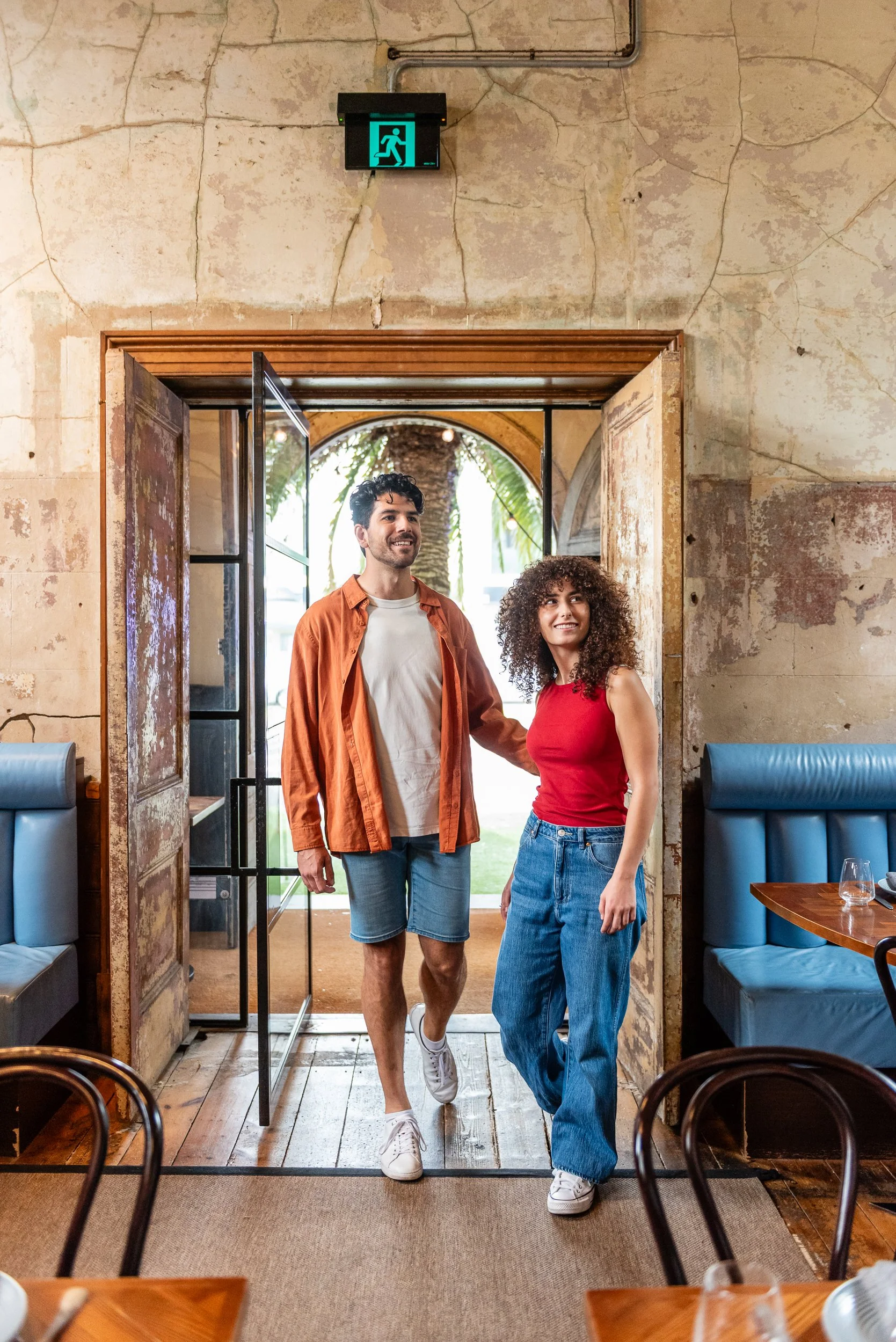 A man and a woman walking out of a restaurant door, smiling. The man has dark hair, a beard, and wears a light shirt with an orange jacket, and denim shorts. The woman has curly hair, wears a red top and wide jeans. The restaurant has rustic walls, b