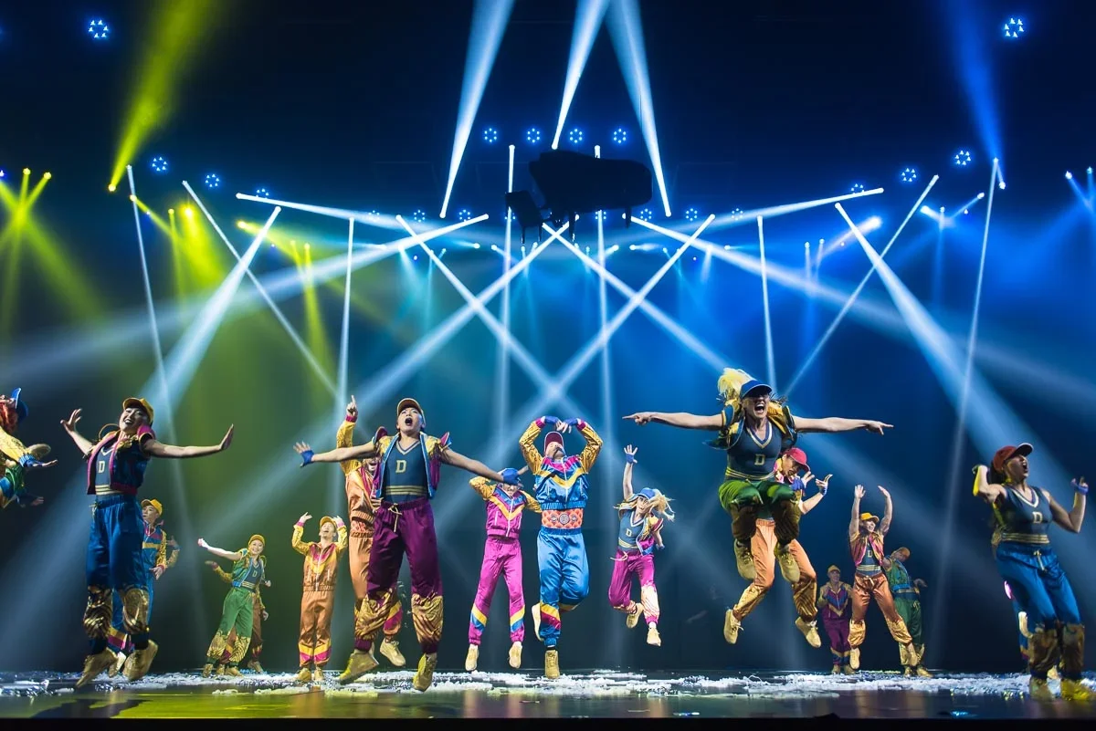 A group of performers on stage with colorful costumes and jumping in mid-air, under bright stage lights and beams, during a lively dance performance.