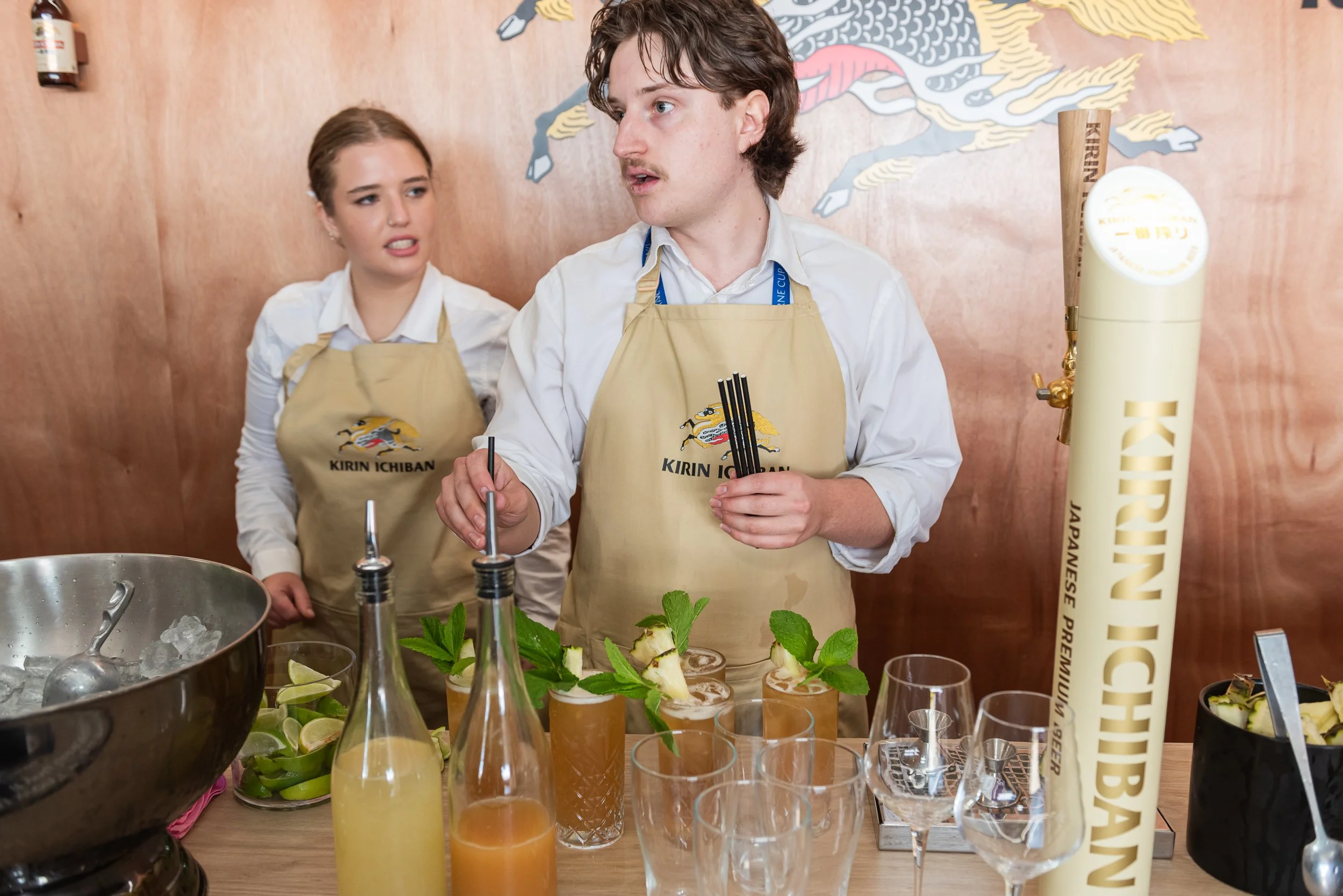 Two bartenders wearing beige aprons with the Kirin Ichiban logo are preparing cocktails behind a bar. One male and one female are visible, with the male actively making drinks. The bar has various bottles, fresh herbs, lime wedges, and glasses, with 