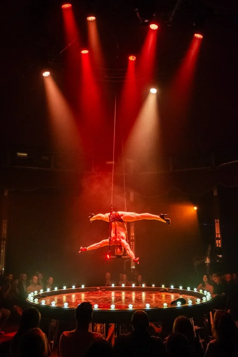 Aerial acrobat performing on a suspended ring in a circus with red and white spotlights, surrounded by an audience