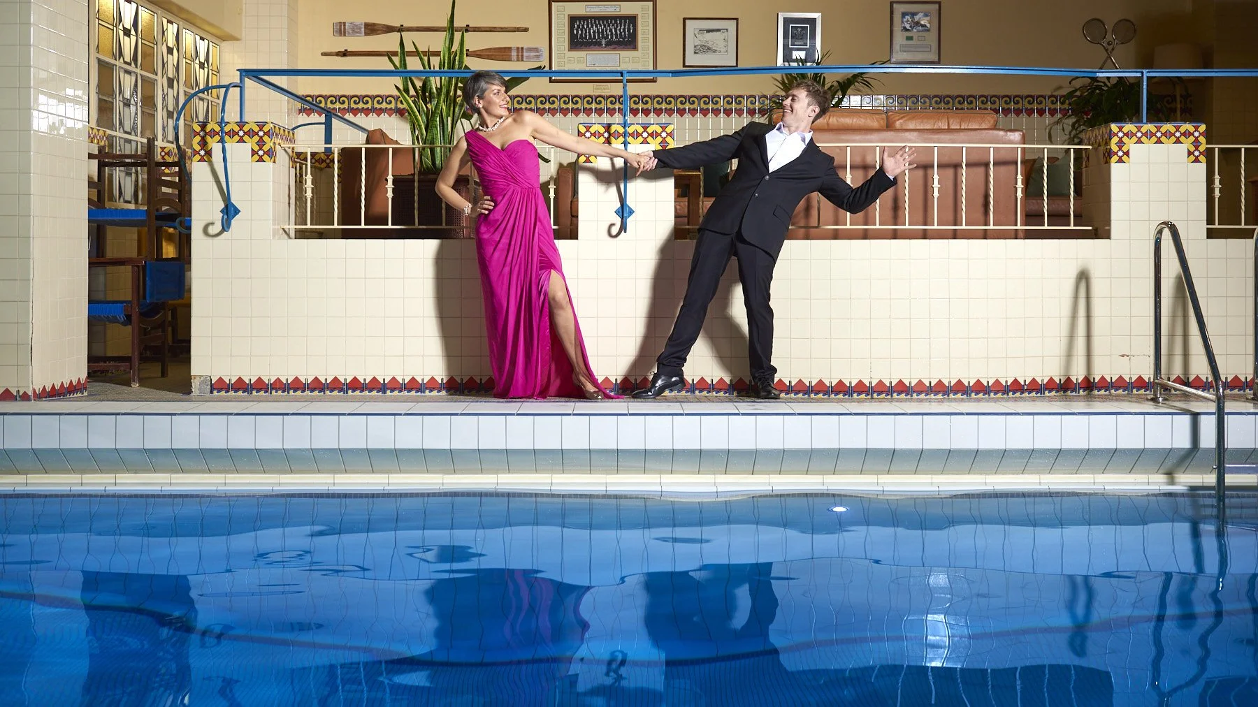 A woman in a pink dress and a man in a black tuxedo are at an indoor pool, holding hands, and seemingly dancing or playing. The woman has one hand on her hip, and the man is leaning back with one hand raised. The setting appears to be a decorated poo