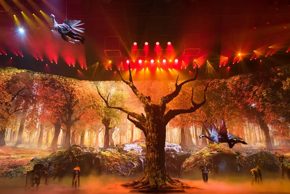 Stage setup with autumn forest scene, large central tree, hanging puppet bird, palm like wings, and human puppeteers with fog at the floor, illuminated with red and yellow stage lights.