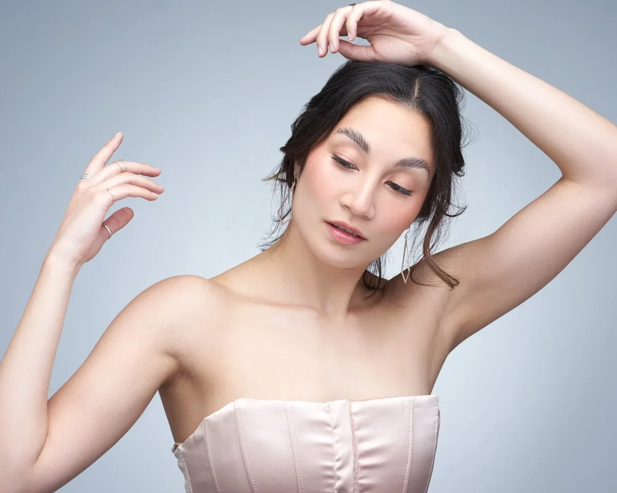 A woman with dark hair and light makeup, wearing a strapless satin top, poses with her eyes closed and one hand above her head, against a light gray background.