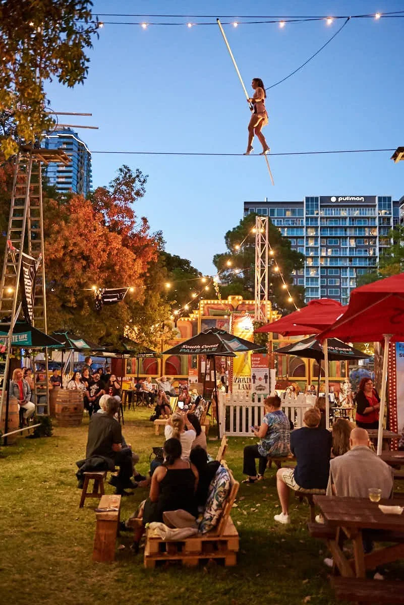 A young girl walking on tightrope high above a fairground, holding a balancing pole, with spectators watching from the ground.