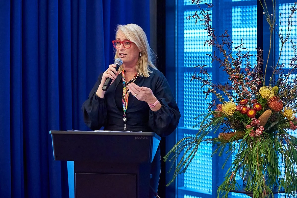 A woman with blonde hair, red glasses, wearing a dark jacket, standing at a podium and speaking into a microphone, with a large colorful floral arrangement beside her and a blue curtained backdrop.