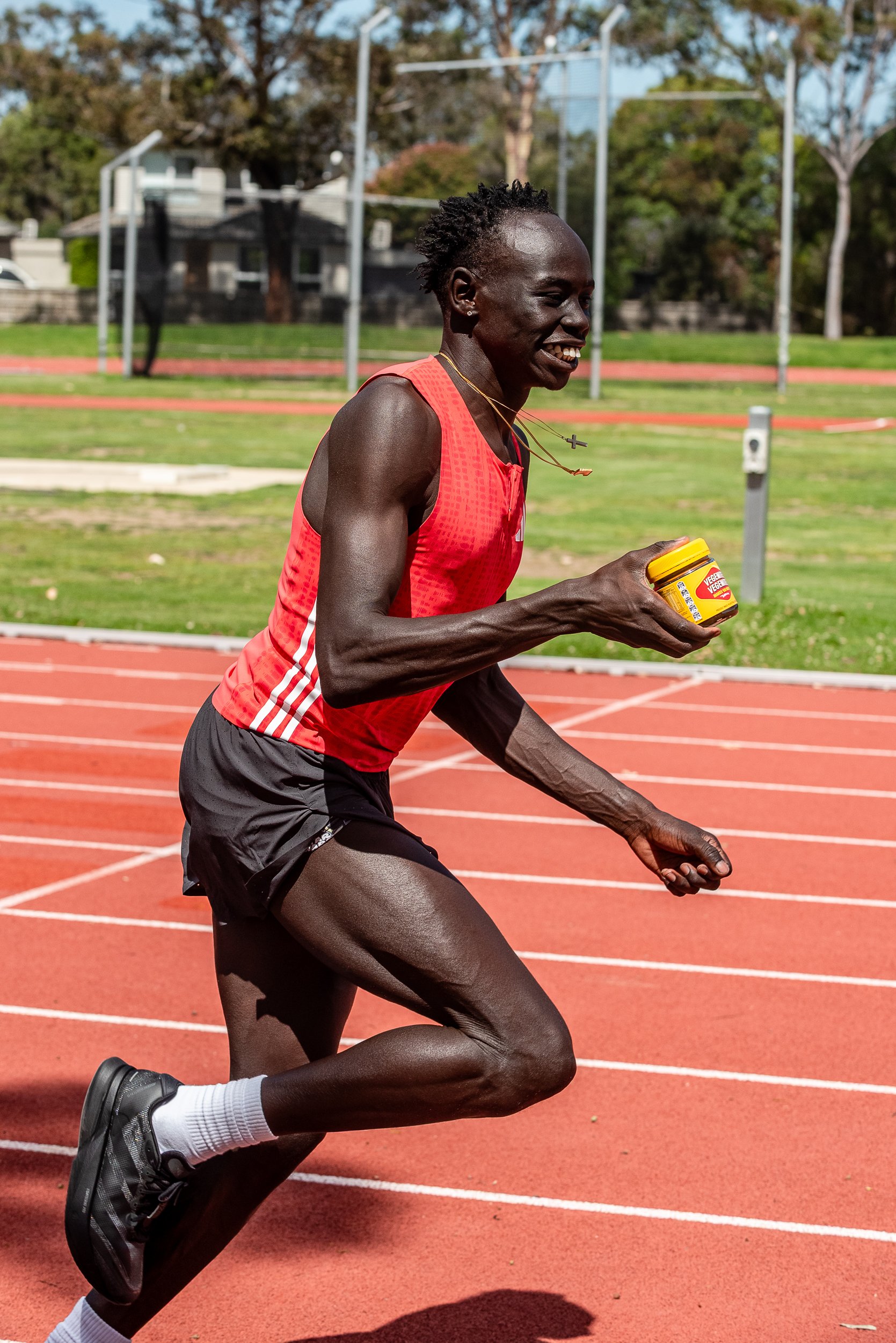 A male runner in a red athletic tank top and black shorts running on a red track with a smile, holding a yellow container of mustard, with a green field and trees in the background.