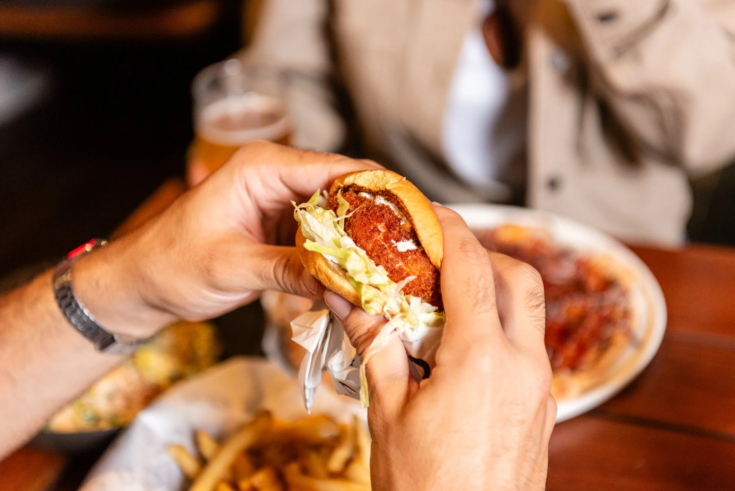 A person holding a small sandwich with lettuce, crispy meat, and sauce, with a plate of similar sandwiches in the background.