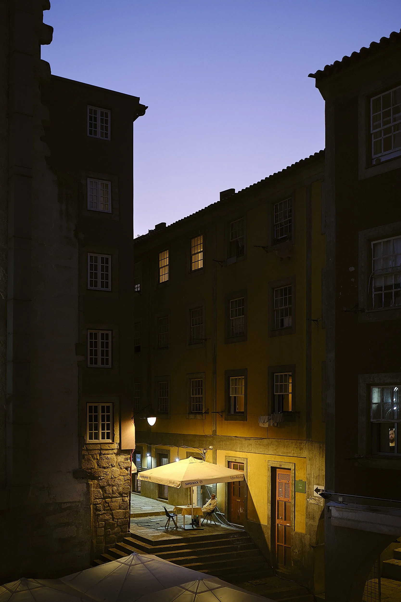 A dimly lit outdoor cafe in a narrow alleyway at dusk, with a person sitting under a large umbrella near a yellow building.
