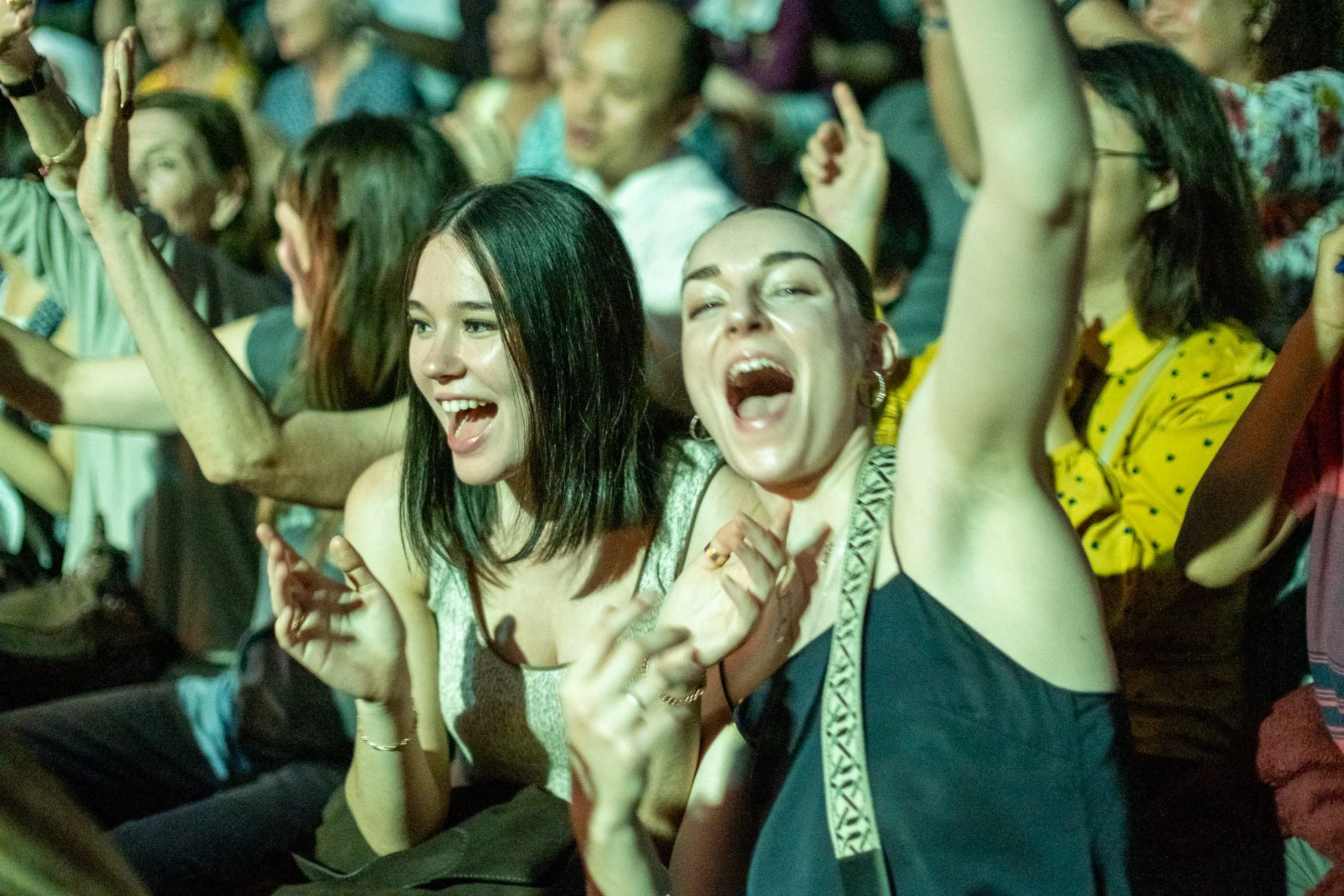 Two women in the front are smiling and singing, one with her eyes closed and the other with her mouth open wide, at a crowded event or concert. The woman on the left has straight, dark hair and is wearing a sleeveless top; the woman on the right has 