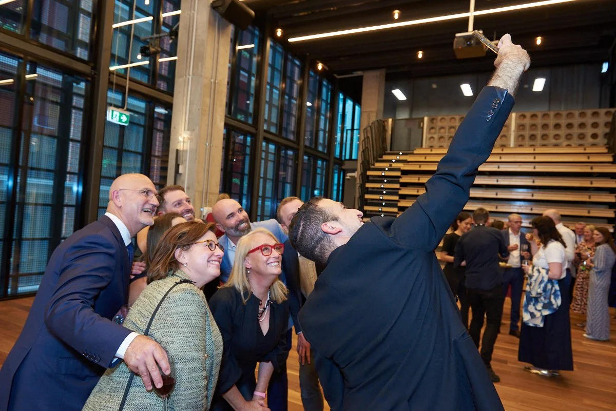 Man taking a selfie with a group of laughing people at a social gathering in a modern indoor space.