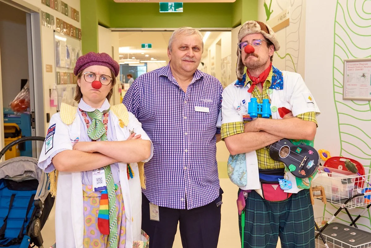 Two adults and one person dressed as a clown in a hospital corridor. The two dressed as clowns are wearing colorful costumes, red noses, and are crossing their arms with serious or grumpy expressions. The man in the middle is in casual checkered shir