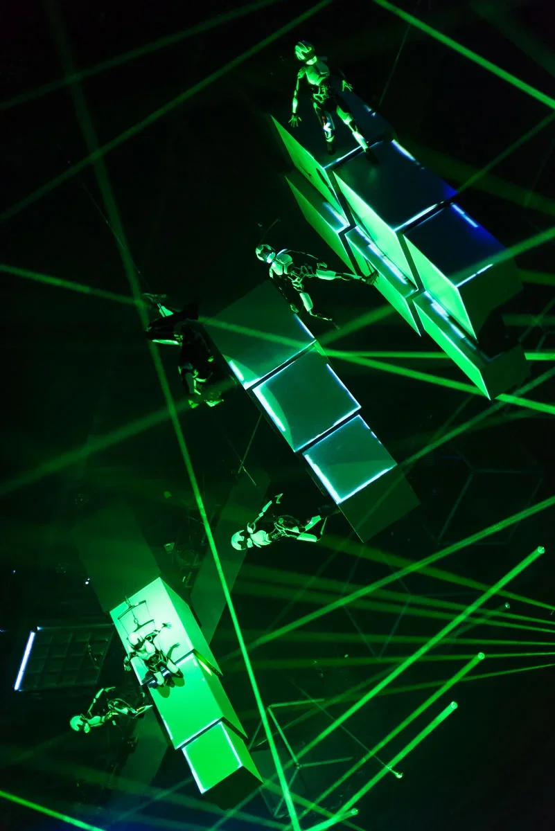 Four people wearing futuristic suits with glowing accents sit on illuminated metallic platforms suspended in midair, surrounded by green laser beams creating a high-tech, sci-fi atmosphere.