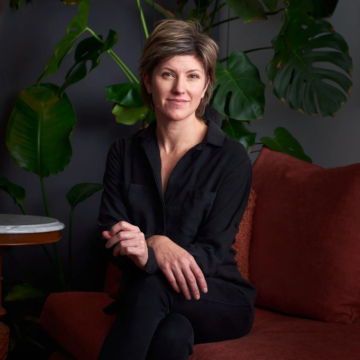 A woman with short brown hair, wearing a black shirt, sitting on a red cushion couch, with large green plants in the background.