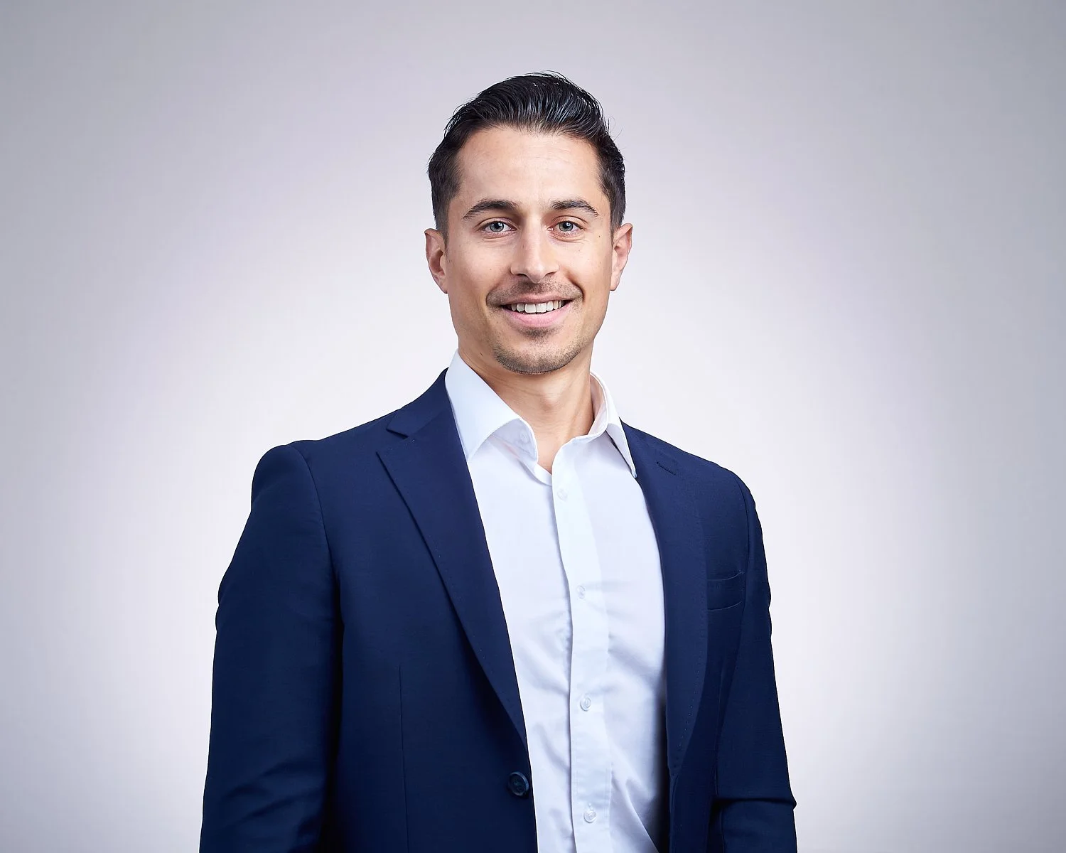 A young man with dark, slicked-back hair, wearing a navy blue suit and white dress shirt, smiling against a plain light gray background.