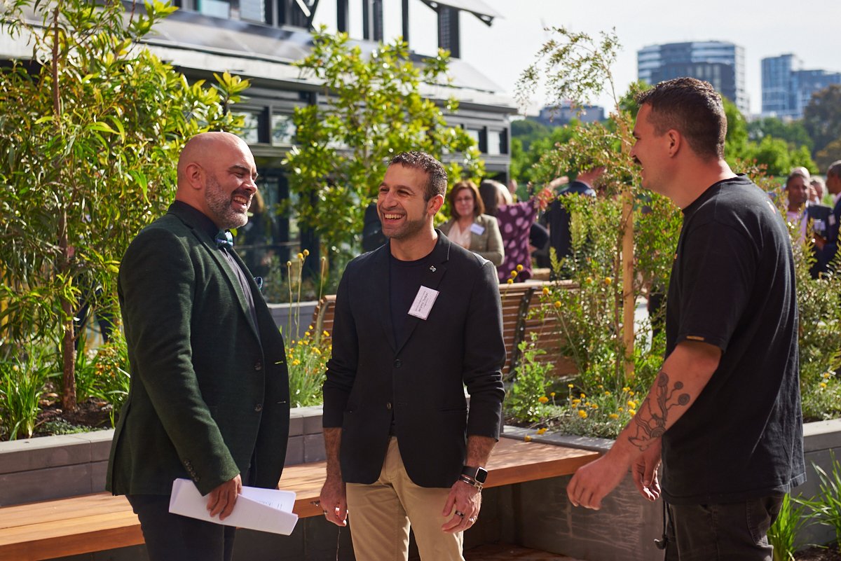 Three men are engaged in conversation at an outdoor event, with greenery and city buildings in the background.