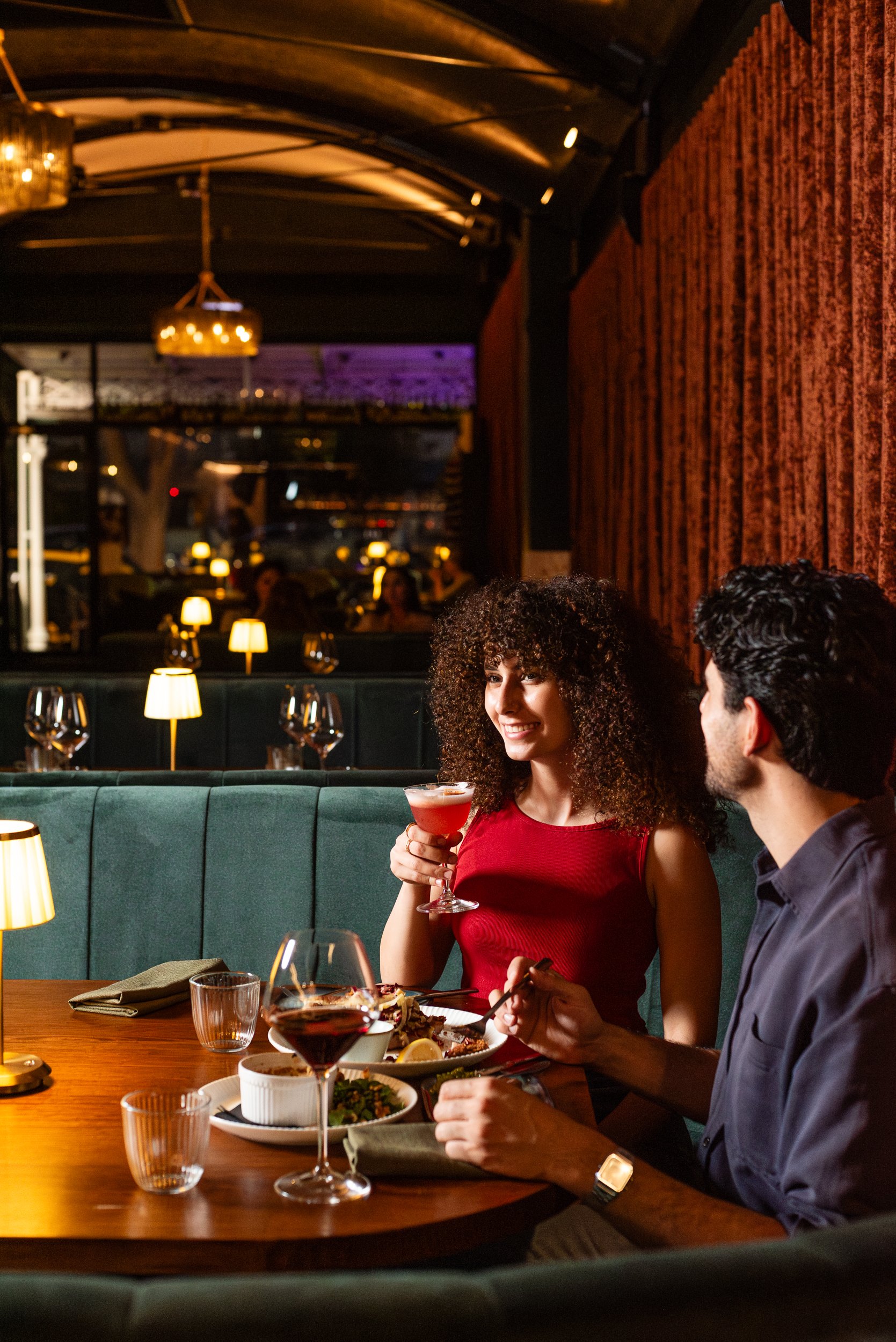 Two people dining in a warmly lit restaurant, one woman holding a cocktail and smiling, the other man with dark hair; table set with food, wine glasses, and tableware. Background features cozy lamps and decor.