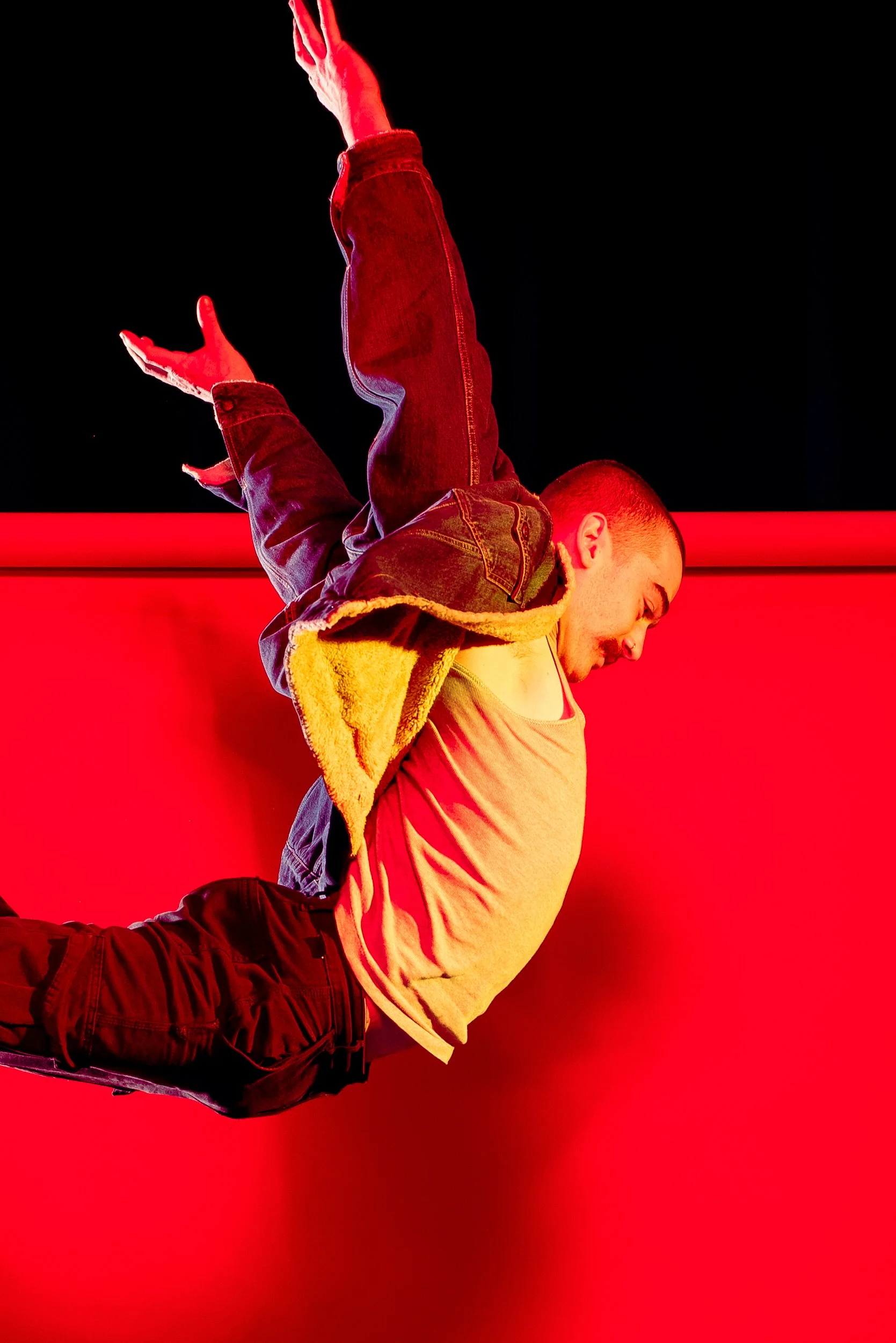 A man with a shaved head wearing a beige tank top, a yellow and brown jacket, and black pants, is leaning back with his arms raised, against a red and black background.