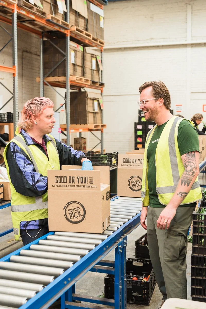 Two workers in a warehouse chatting near a conveyor belt with boxes labeled 'Farmers Pick' and 'Good on You'. The warehouse has tall shelves with stacked boxes.