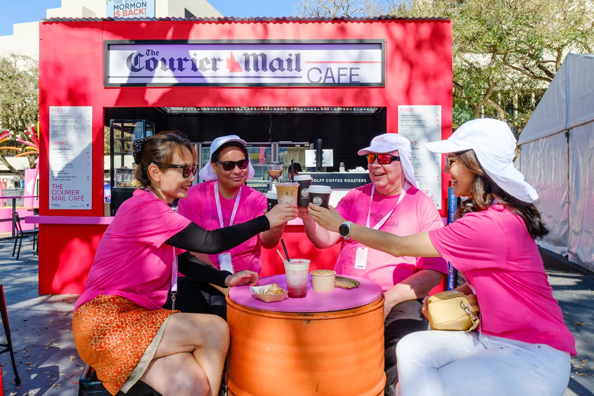 Four women wearing pink t-shirts and sunglasses sit around an orange barrel table at an outdoor event, smiling and clinking drinks, with a food stand labeled 'The Courier Mail Café' behind them.