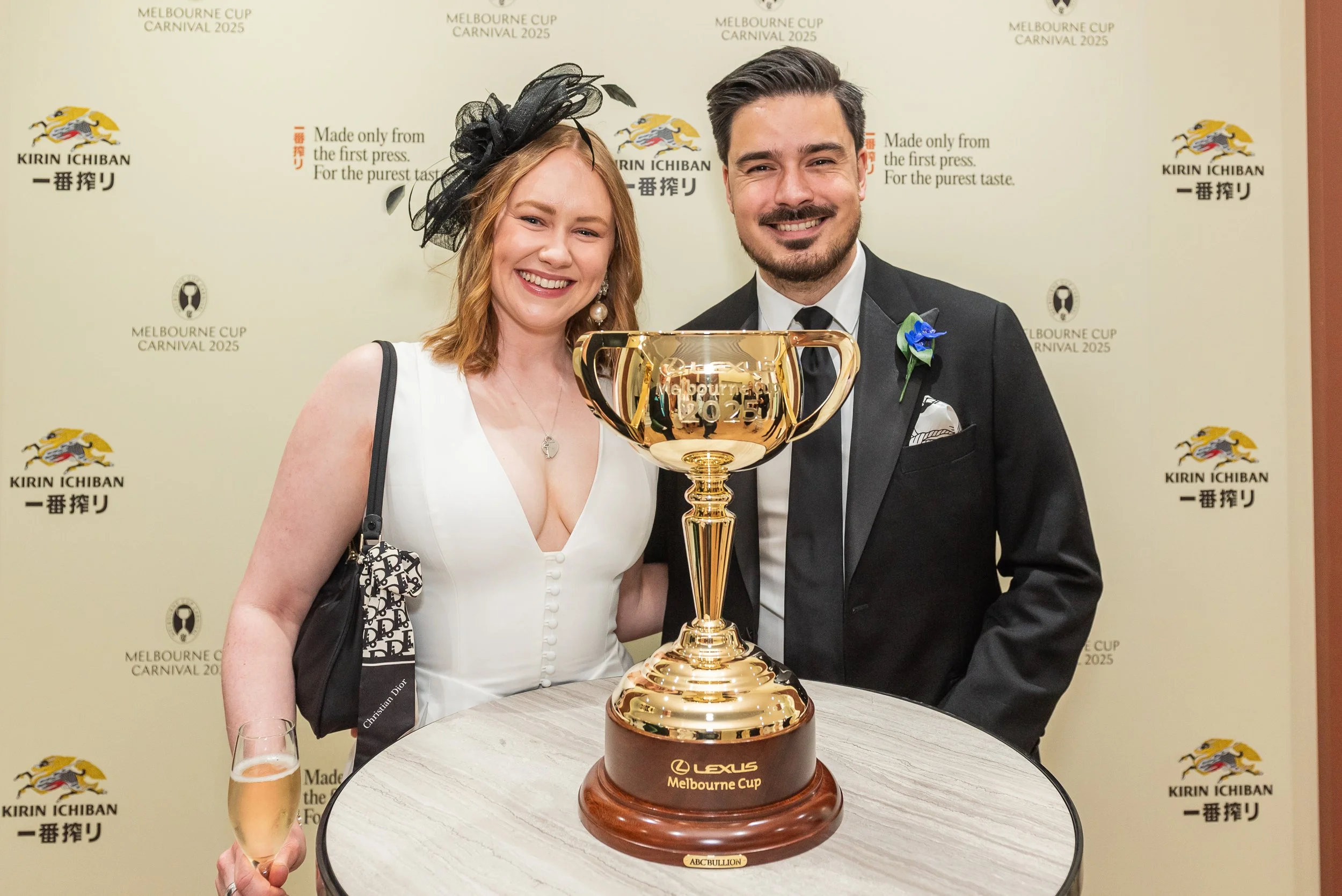 A smiling woman in a white dress and a black hat with a veil stands next to a smiling man in a black tuxedo with a blue flower pinned to his lapel. They are holding a large gold trophy with the words 'Melbourne Cup 2025' engraved on it, on a table in