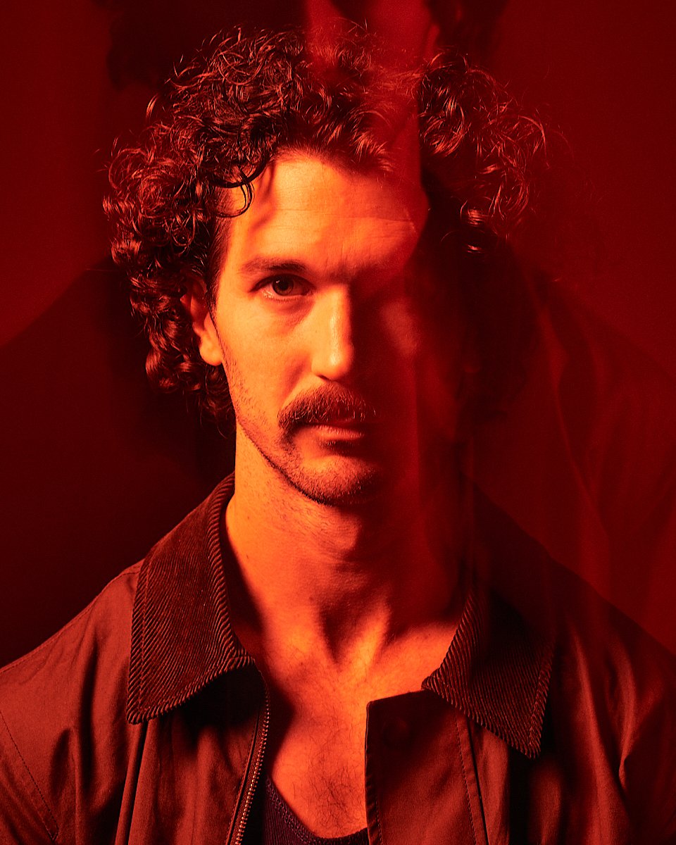 A man with curly hair and a mustache looks directly at the camera. The photo has a red lighting effect, creating a dramatic and intense atmosphere.