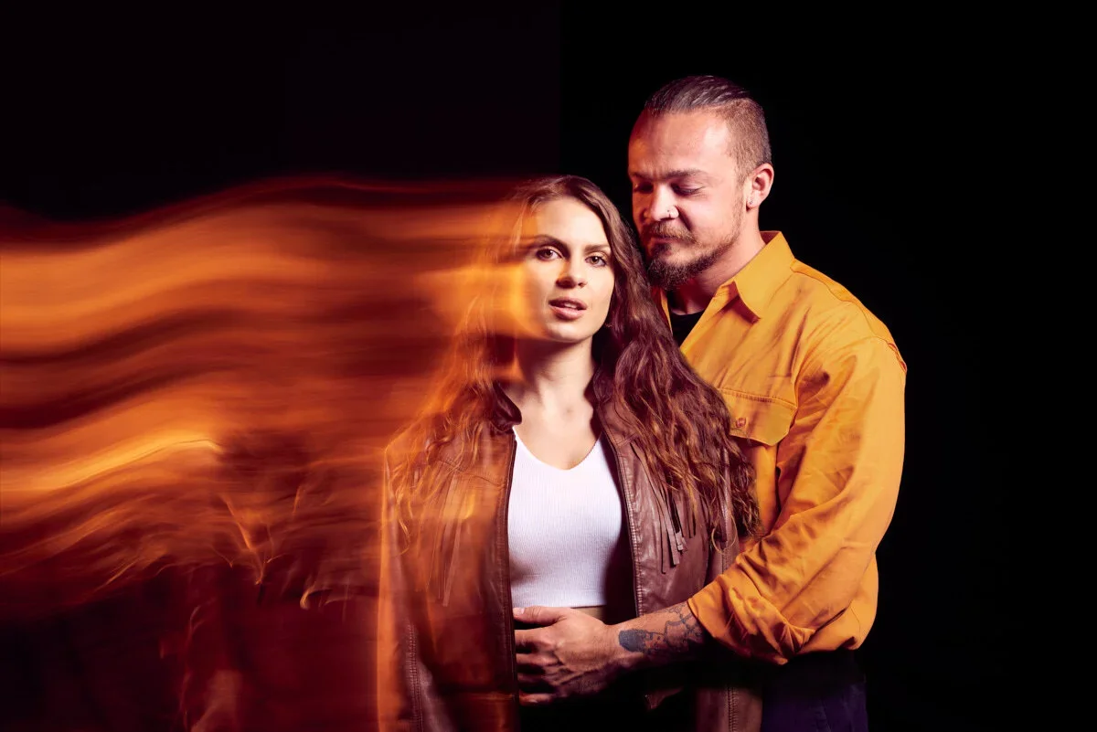 A woman with long, curly hair and a brown leather jacket stands in front of a man with a beard and a yellow shirt, who is holding her around the waist. A streak of orange light appears to pass through the woman, creating a dynamic effect.