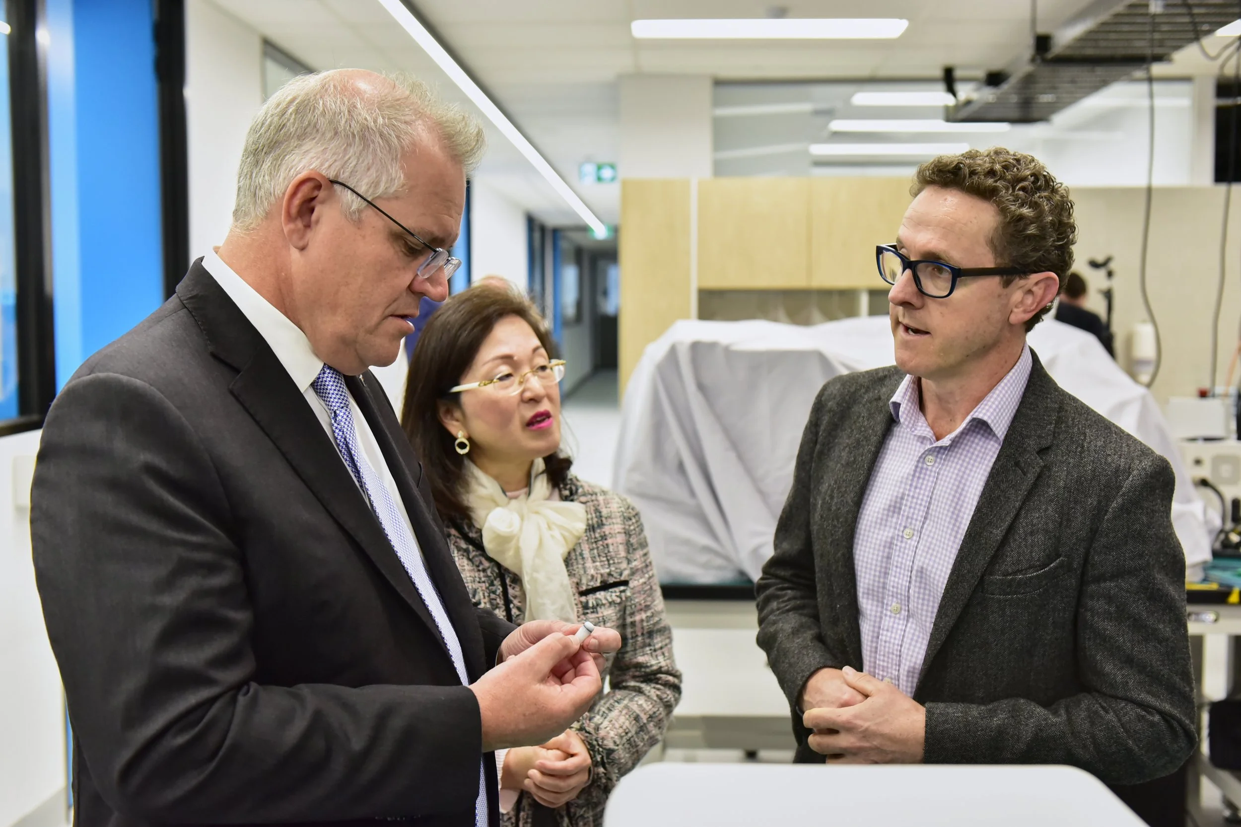 Three people having a discussion in a laboratory, with a man in a suit holding a small object, and a woman and another man listening, in a modern medical or scientific facility.