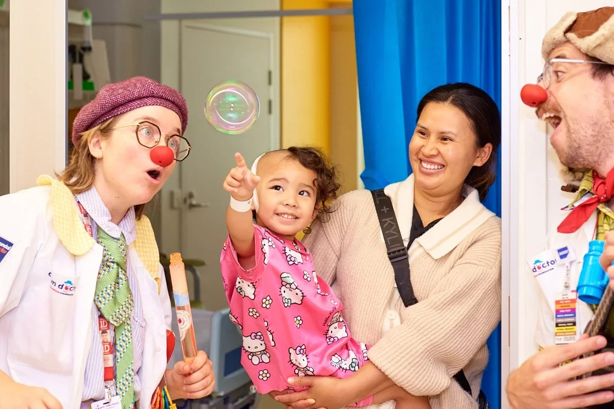 Children and adults, including two dressed as clowns, in a hospital room with a young girl smiling and pointing at a floating soap bubble.