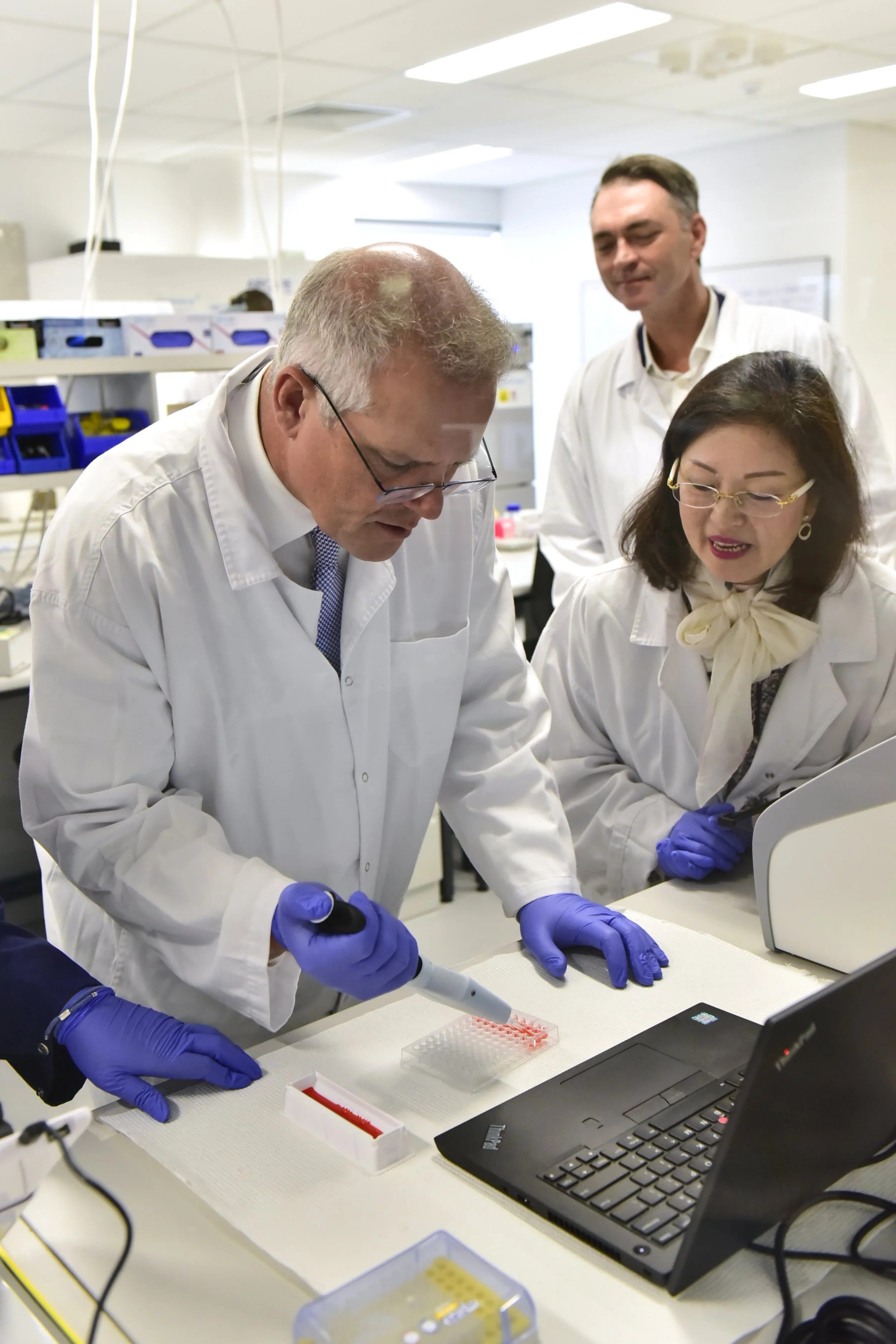 Scientists working in a laboratory, using a pipette to transfer liquid into a well plate, with a laptop nearby.